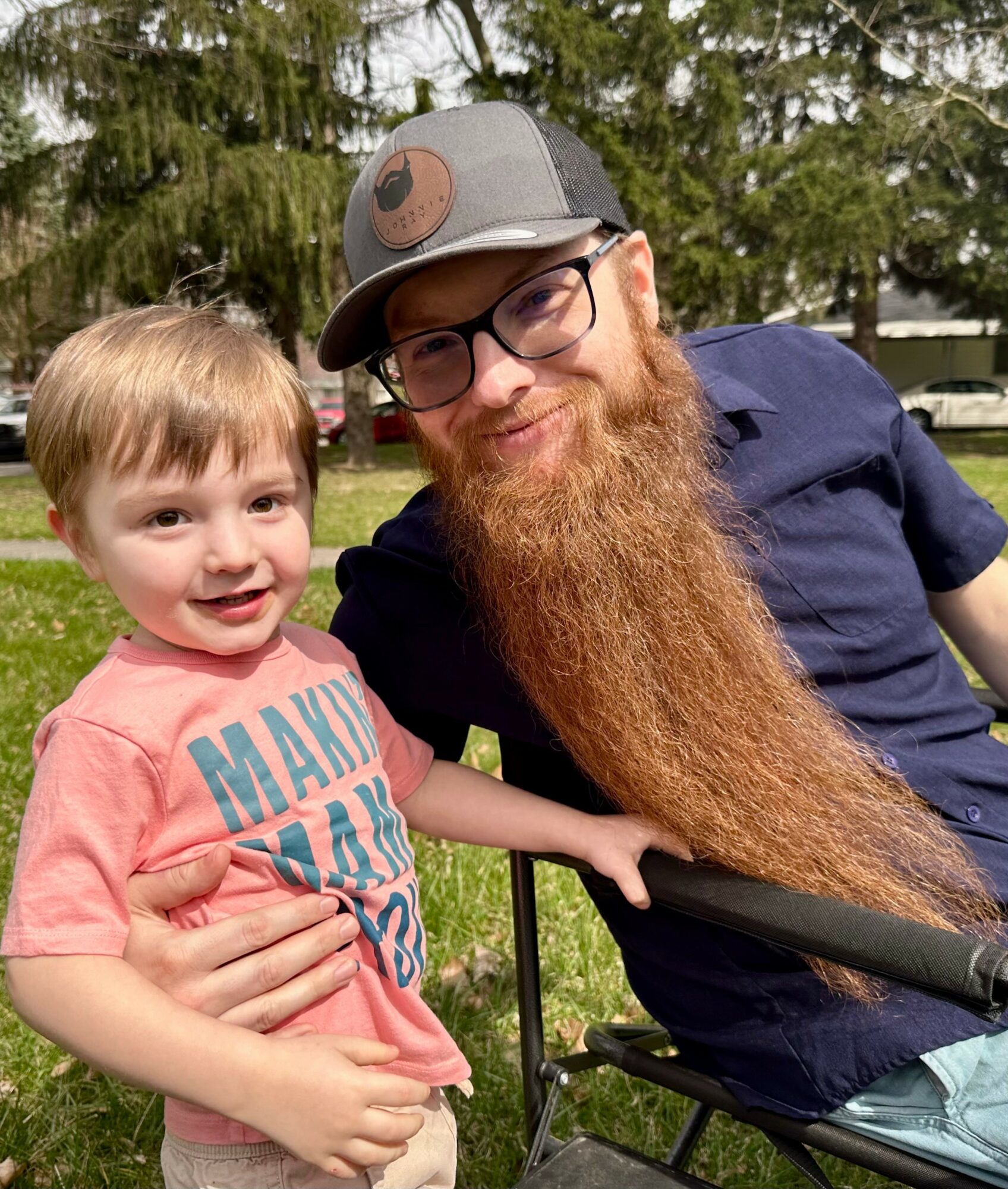 A young boy and a man with a long beard outdoors, smiling, with trees in the background.