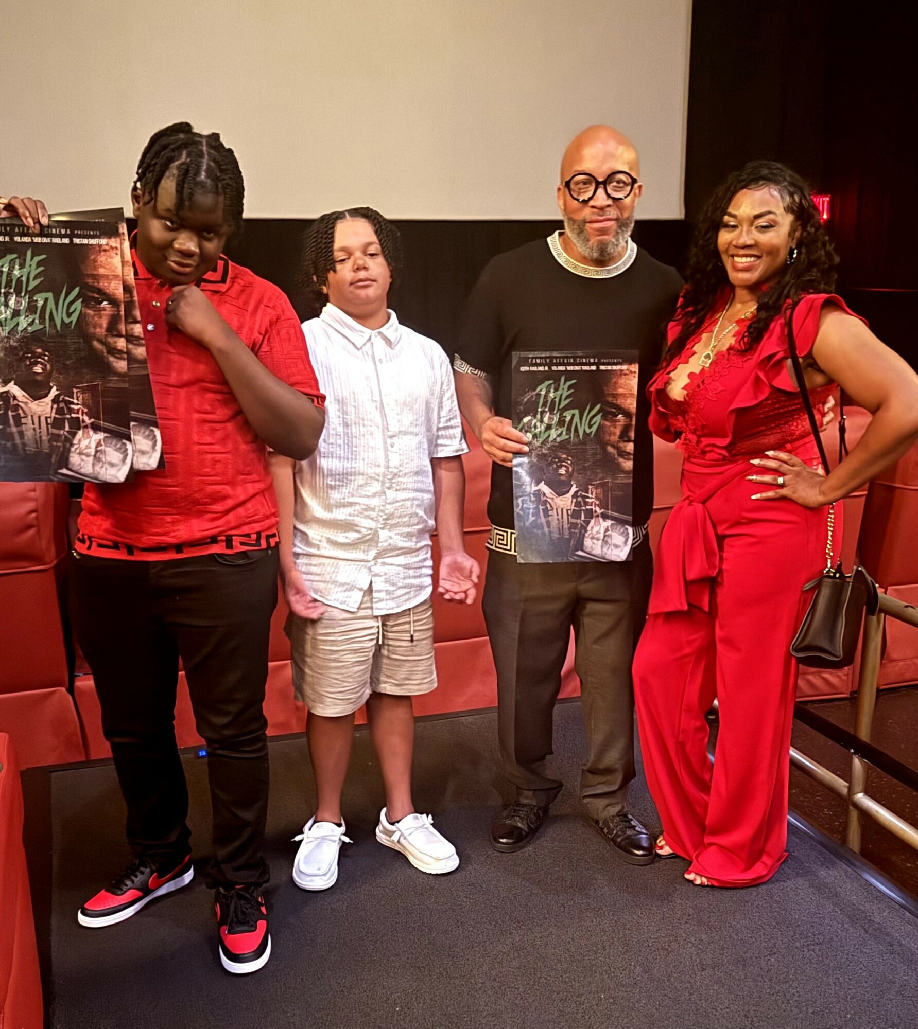 Four people standing indoors, two holding books, with a plain background and red seating. One person wears glasses and a black shirt.