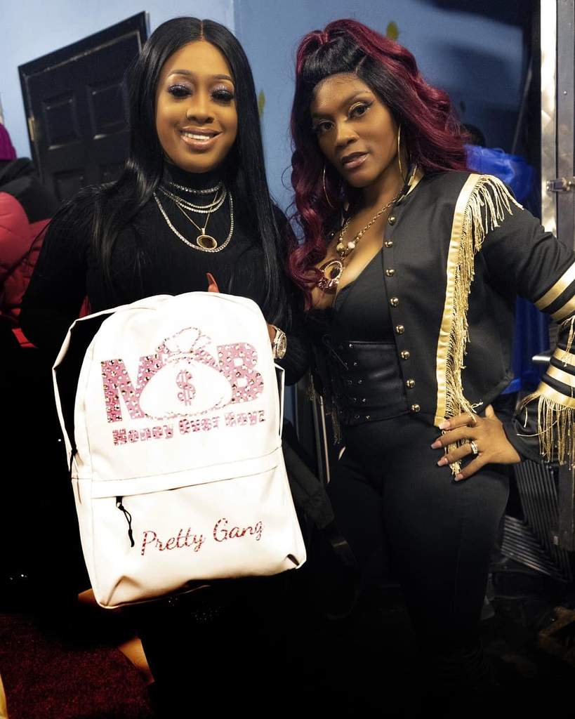 Two women posing indoors, one holding a white bag with pink text, both smiling and dressed stylishly.