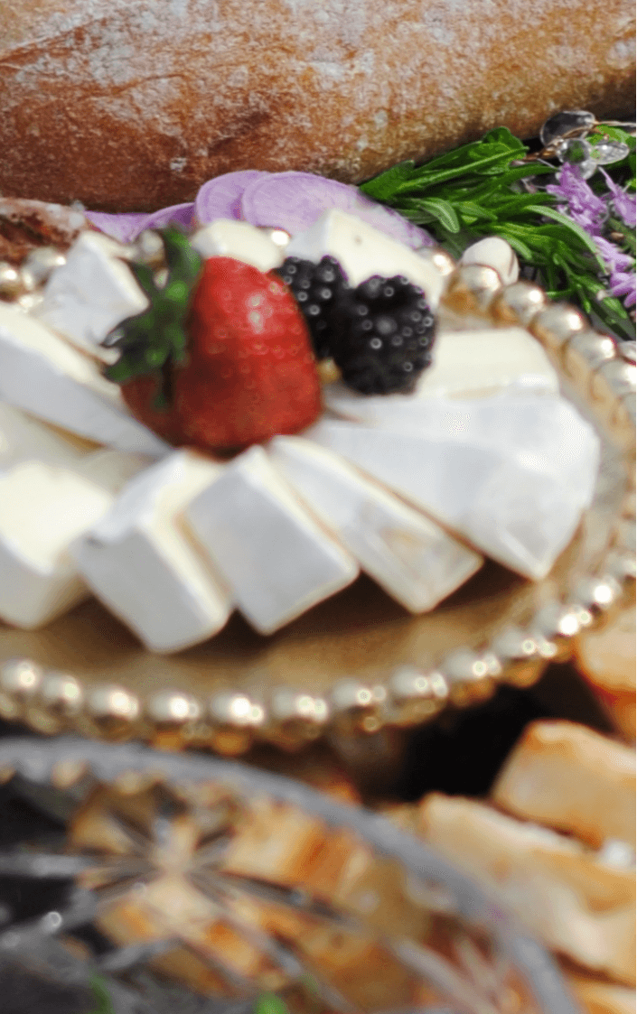 Cheese with strawberries, blackberries, and grapes on a decorative plate, with bread and flowers in the background.