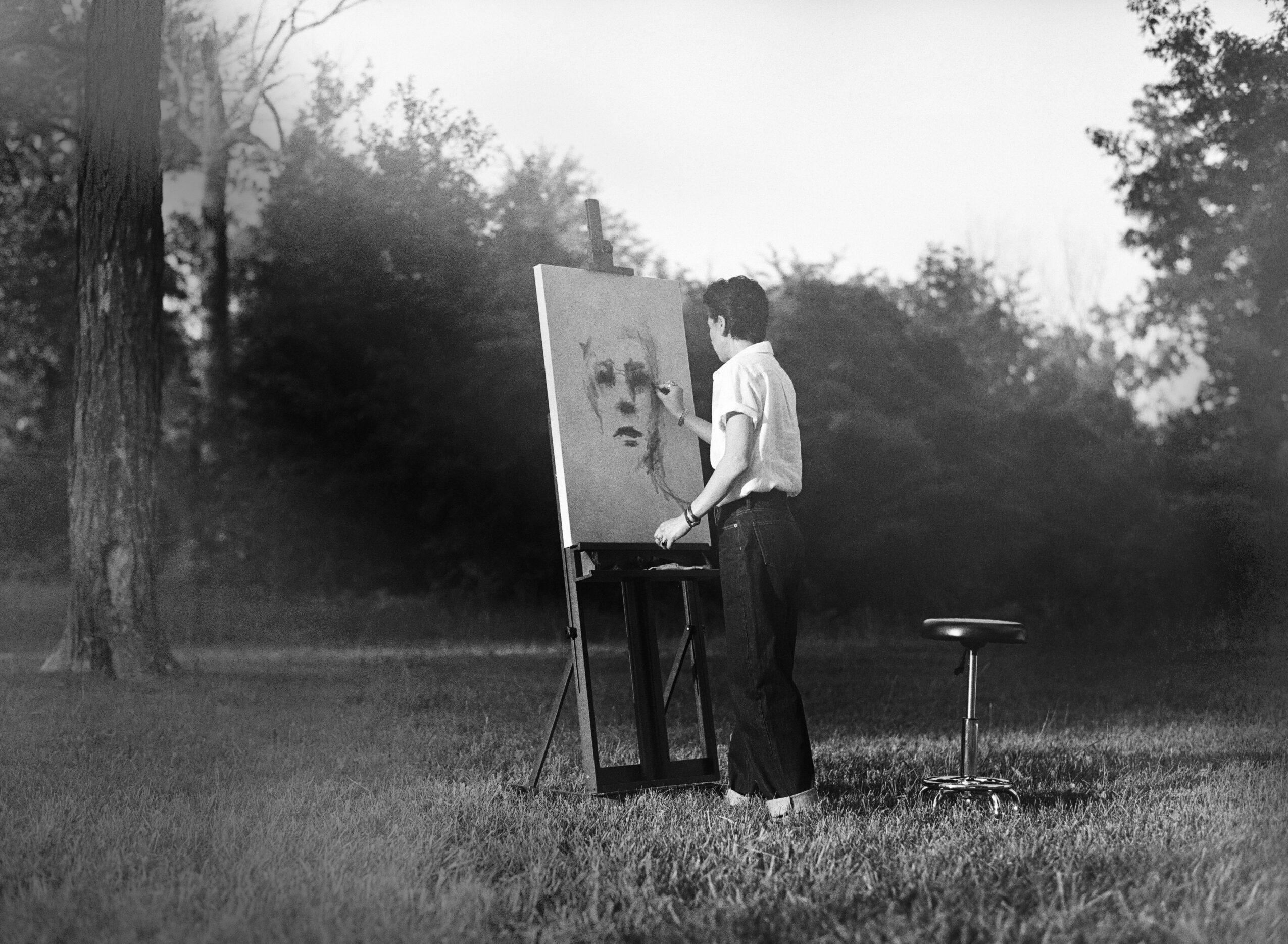 Artist painting a portrait outdoors with trees in the background, standing next to an easel and a stool.