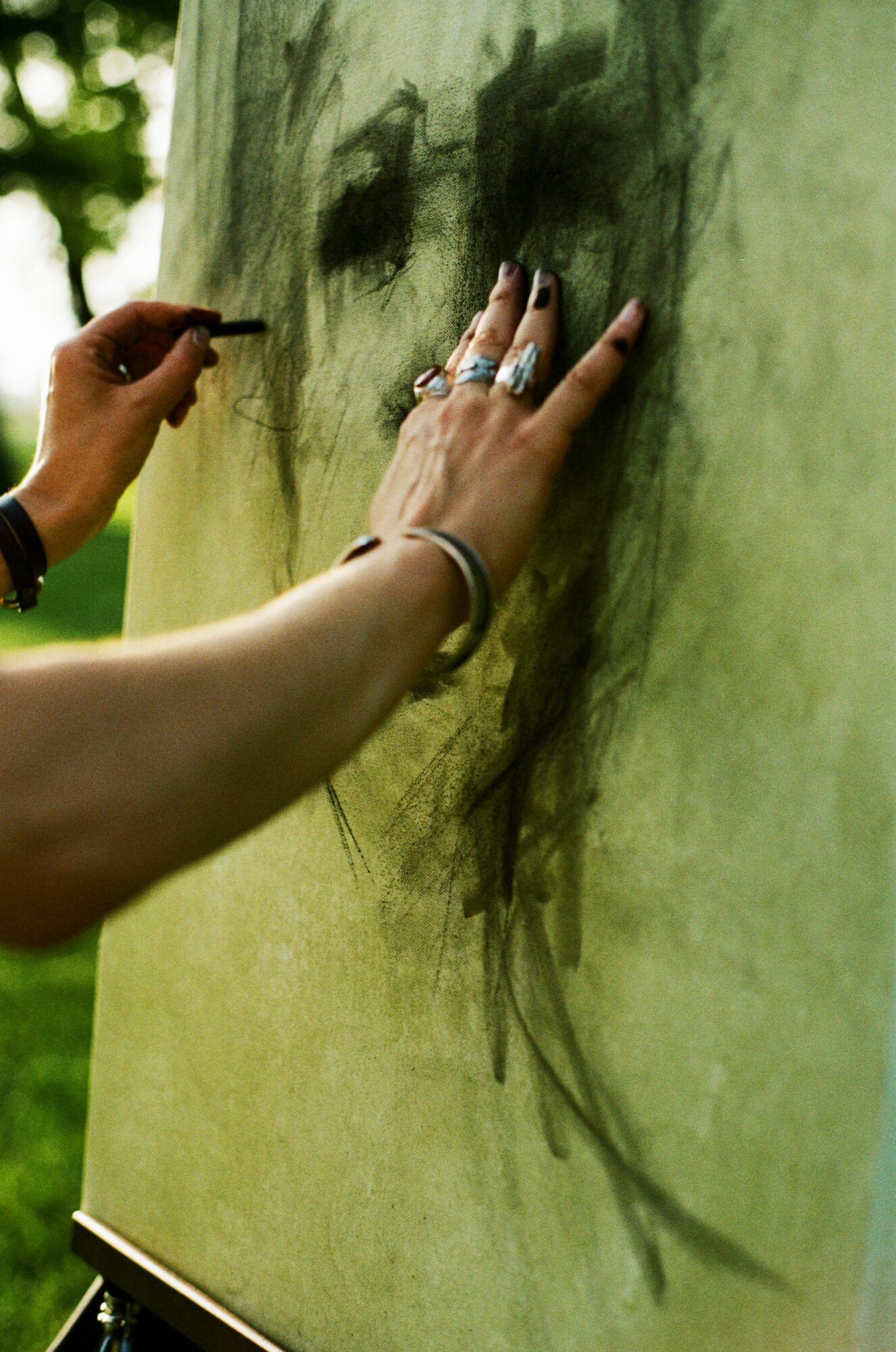 Person drawing on a green tree trunk with charcoal or pencil, wearing rings and bracelets, outdoors.