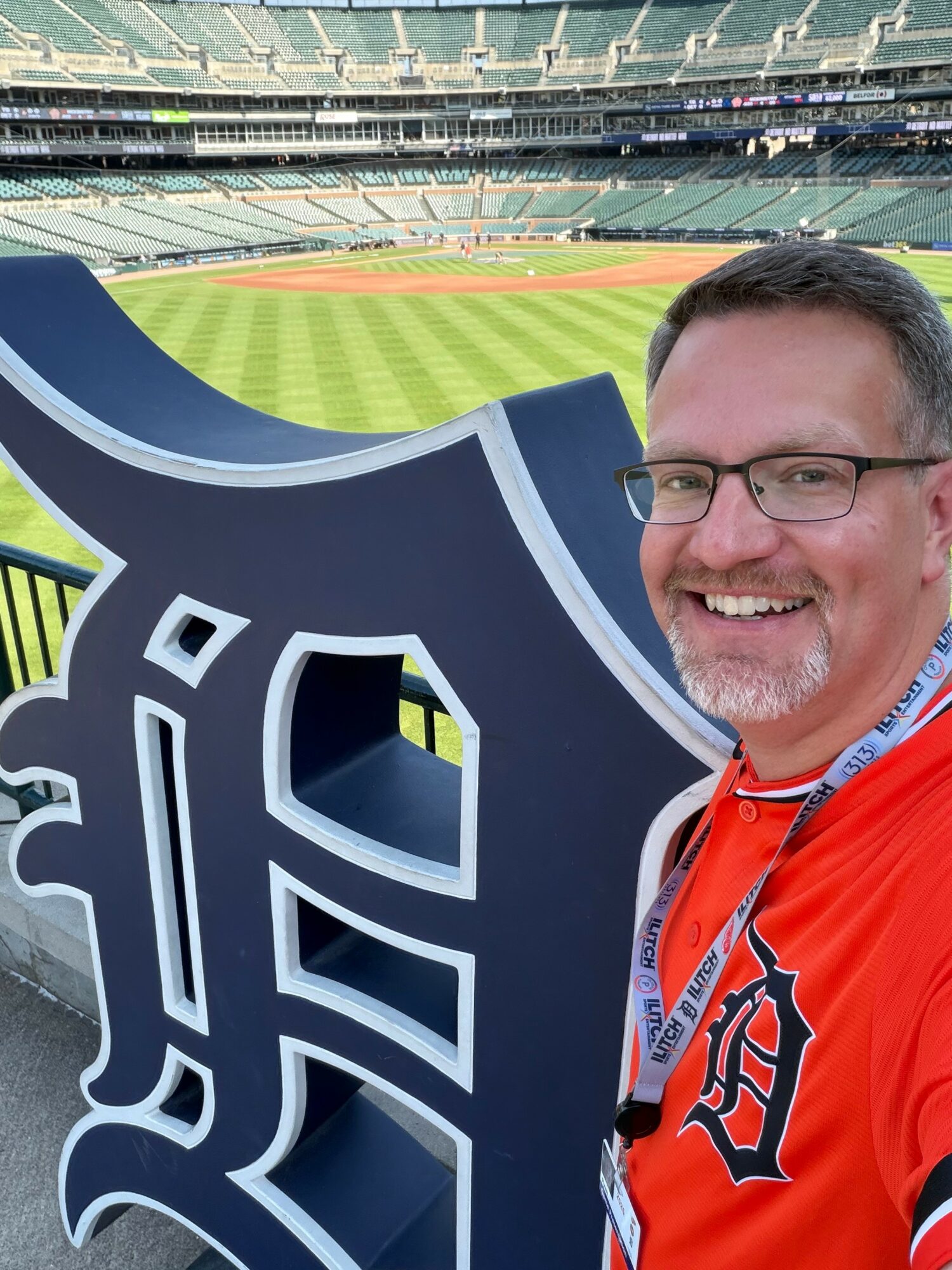 Man with glasses smiling at a baseball stadium, holding a large Detroit Tigers logo, empty seats in background.