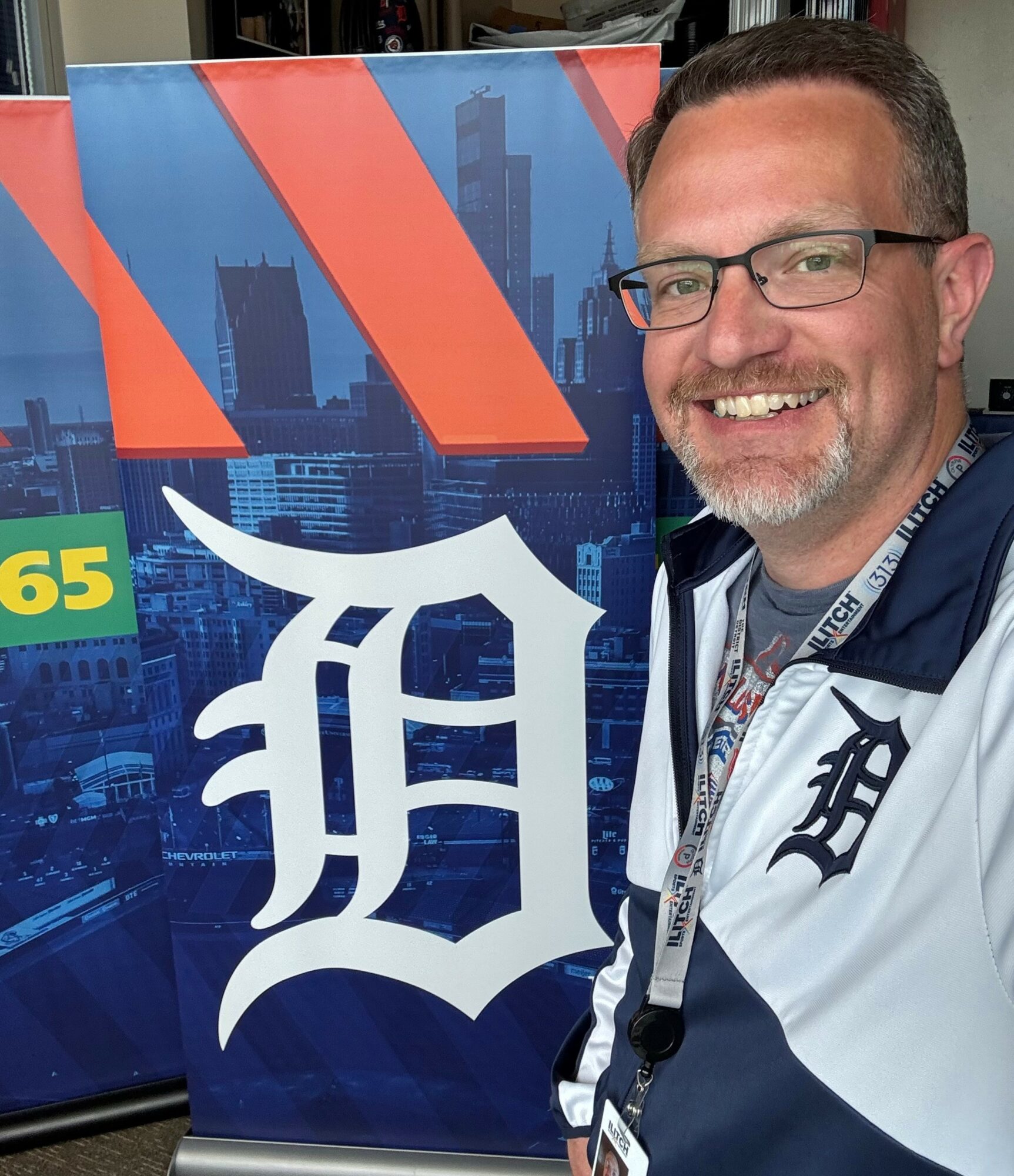 Man with glasses smiling next to a Detroit Tigers banner with city skyline background.