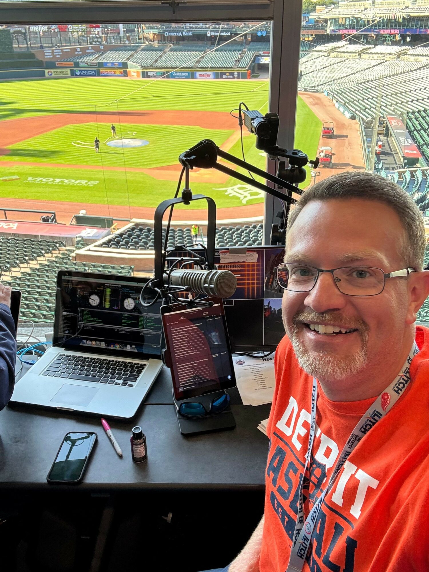 Man smiling in a radio studio with a microphone, laptop, and phone, overlooking a baseball stadium outside the window.