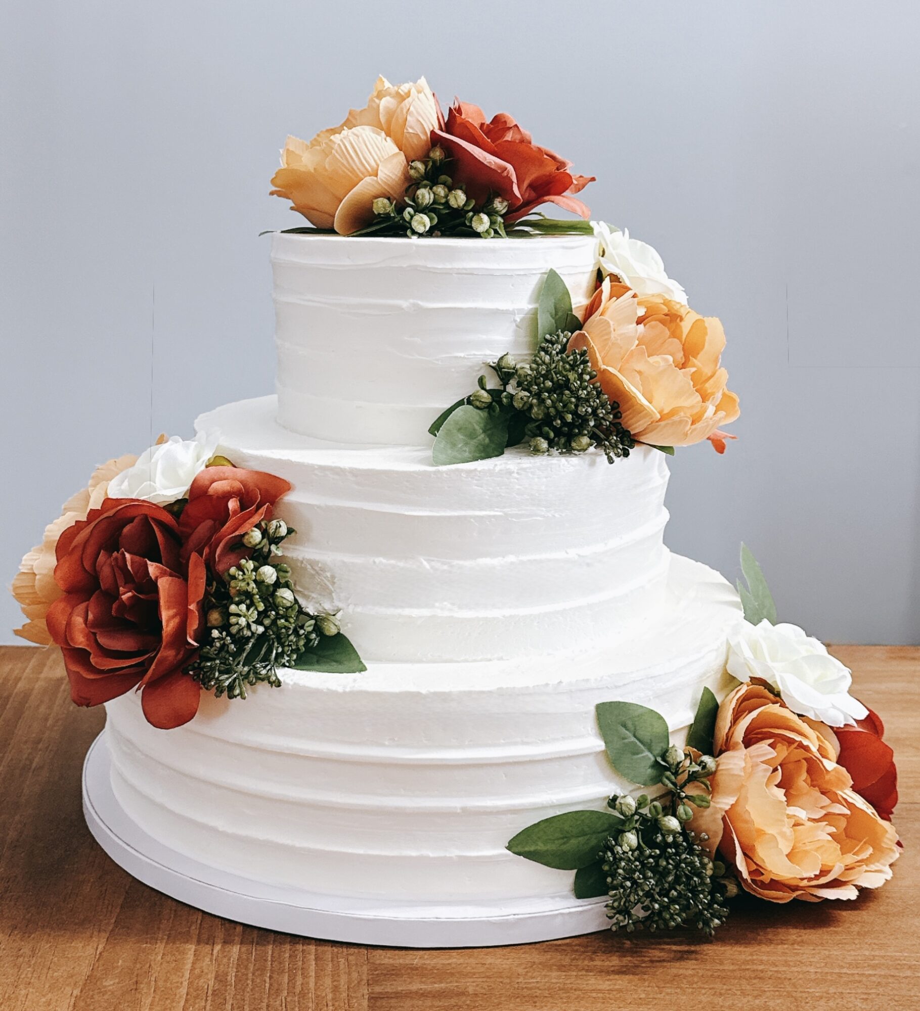 Three-tiered white wedding cake decorated with red and peach roses, green leaves, and small white flowers, on a wooden surface.