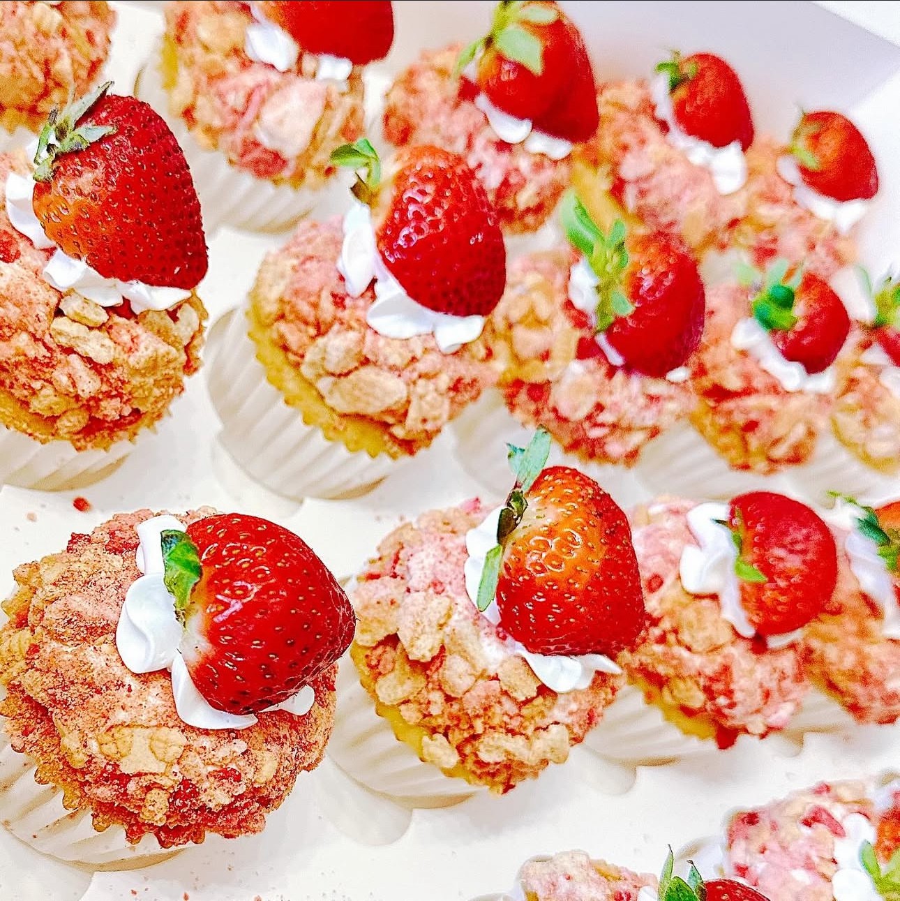 Close-up of strawberry-topped cupcakes with pink crumb topping and white frosting, arranged on a white surface.