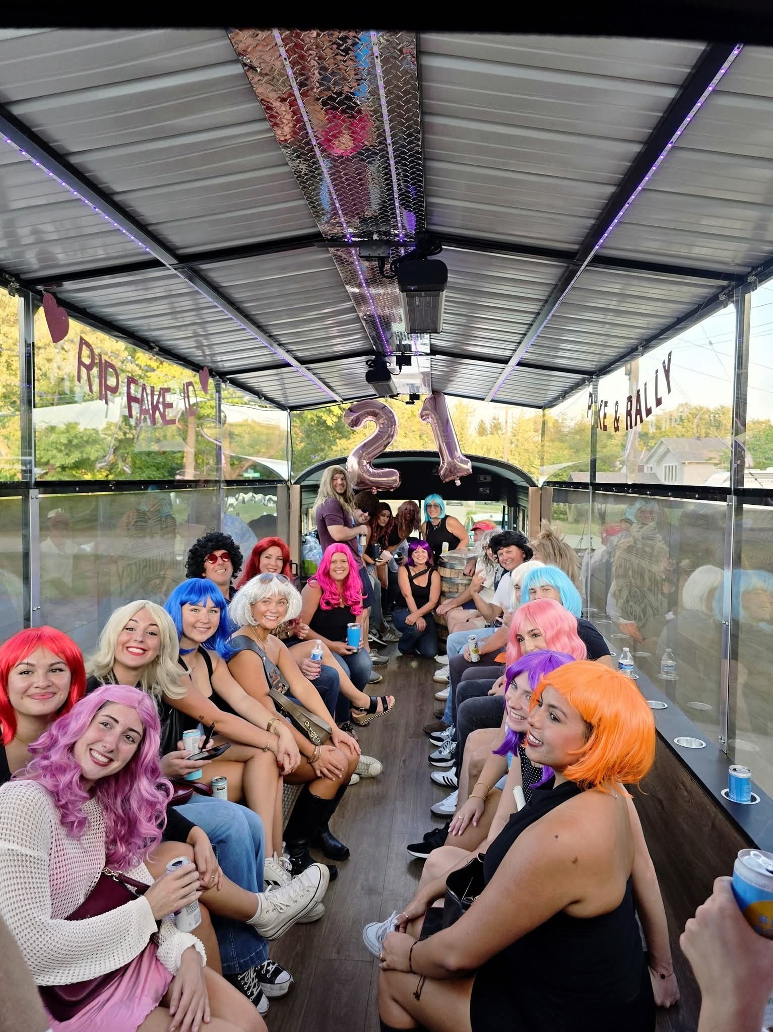 Group of people with colorful hair and costumes sitting inside a glass-enclosed vehicle, celebrating a birthday with balloons and decorations.