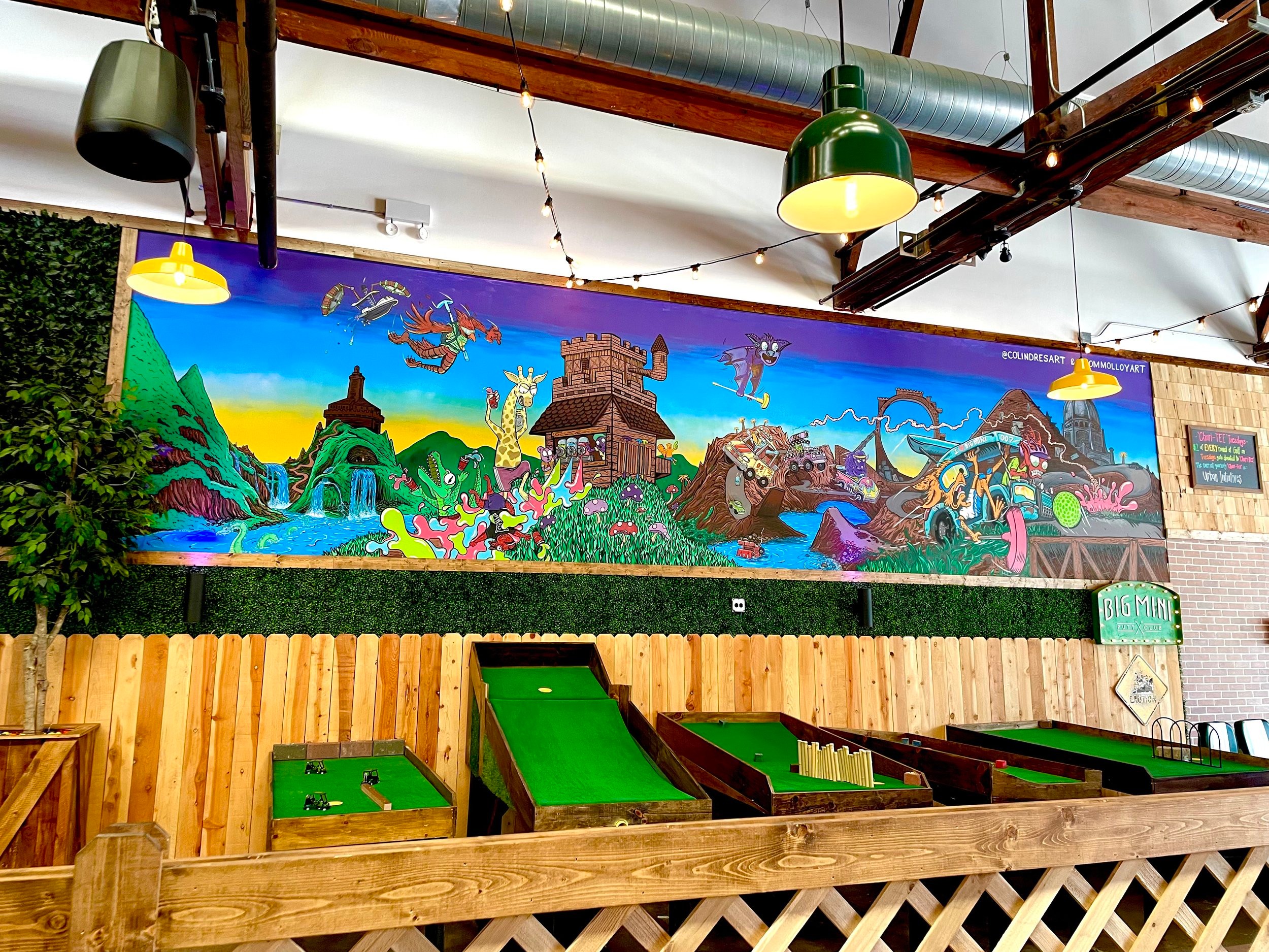 Colorful mural depicting fantasy creatures and landscapes, with a wooden fence and green ramps in front, inside a building with hanging lights.