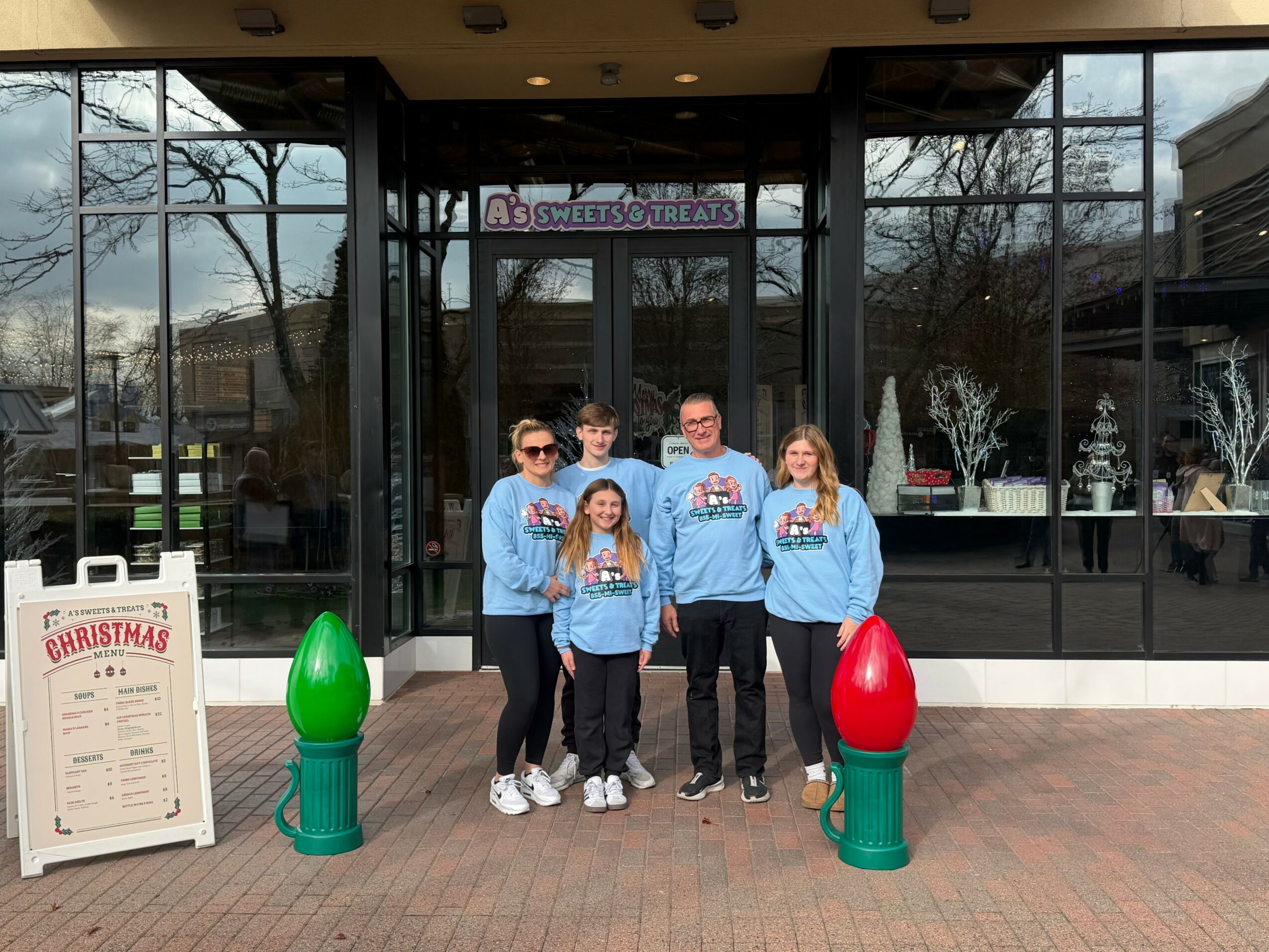 Group of five people standing outside a building with glass doors, two large Christmas decorations, and a signboard, during daytime.