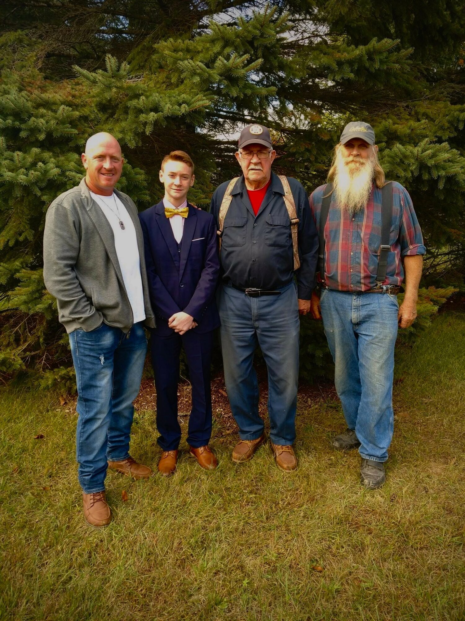 Four men standing outdoors in front of a large tree, dressed casually, with grass beneath them.