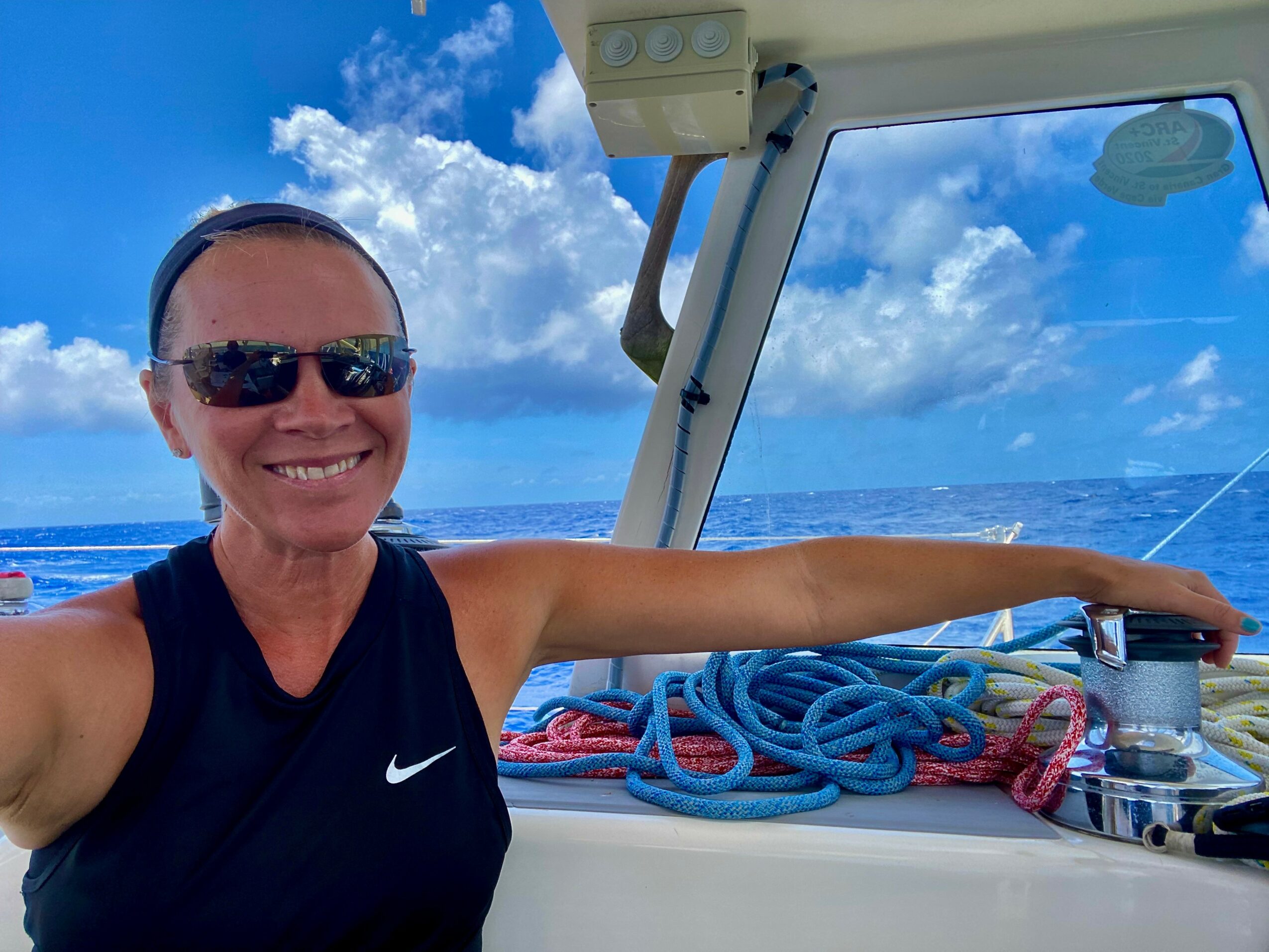 Woman smiling wearing sunglasses on boat with blue sky and ocean background, arm extended, ropes and equipment nearby.