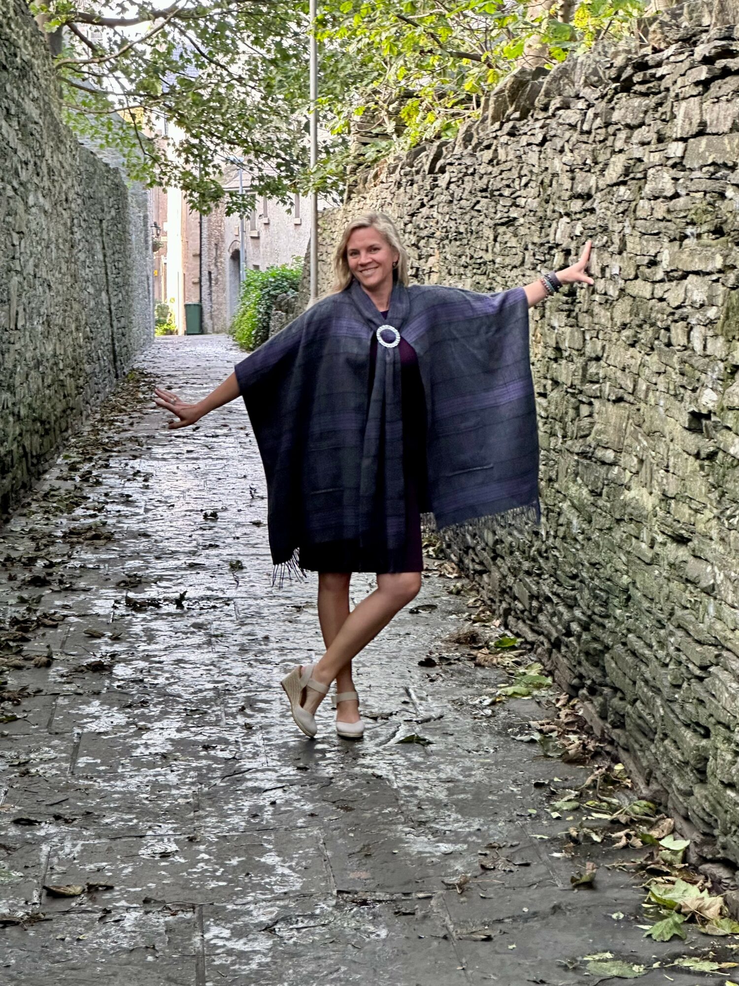 Woman standing on a narrow, wet alleyway with stone walls, arms outstretched, smiling, wearing a dark shawl and white shoes.