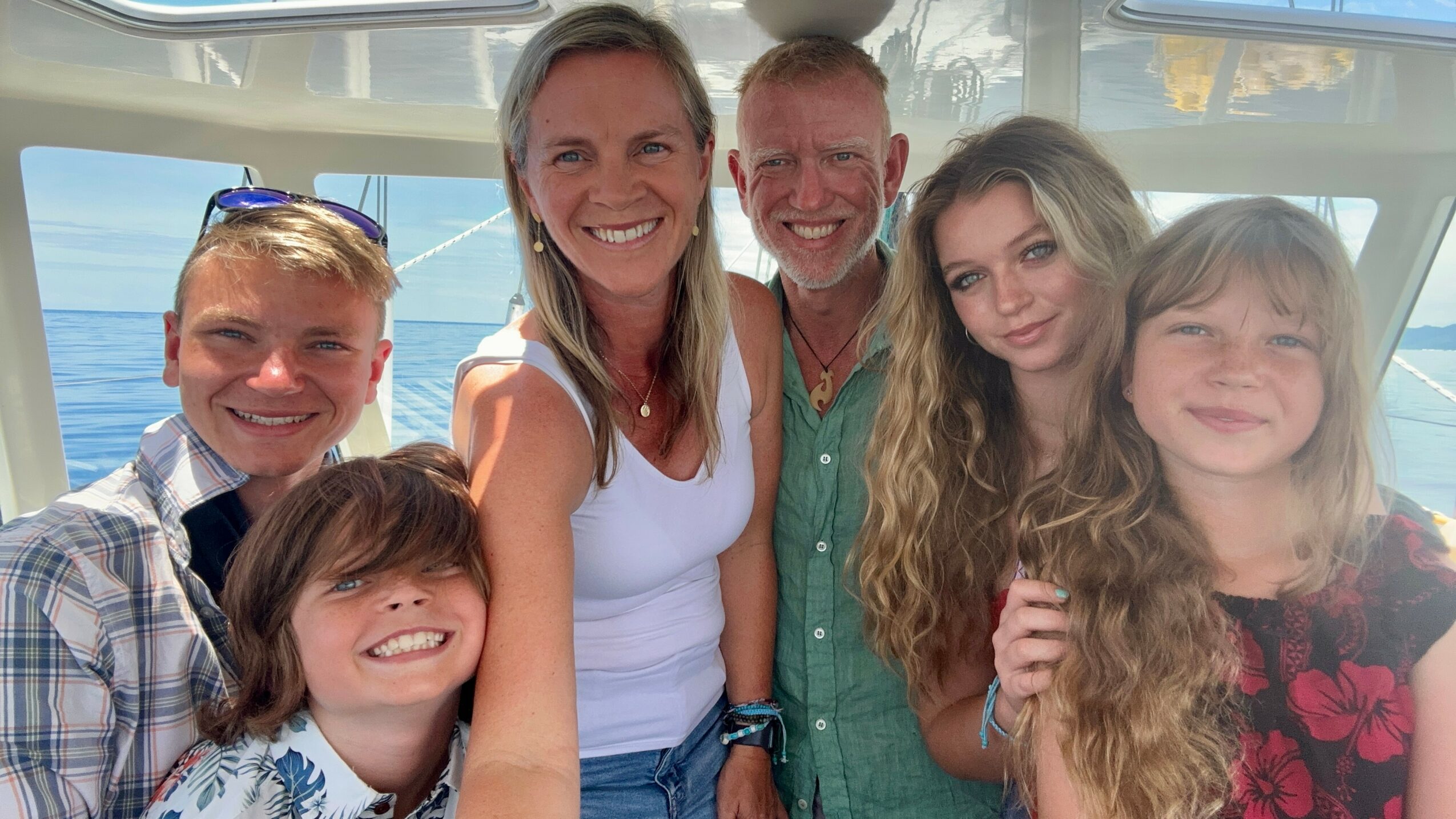 Group of six people smiling on a boat with water in the background, sunlight shining, casual clothing, diverse ages and genders.