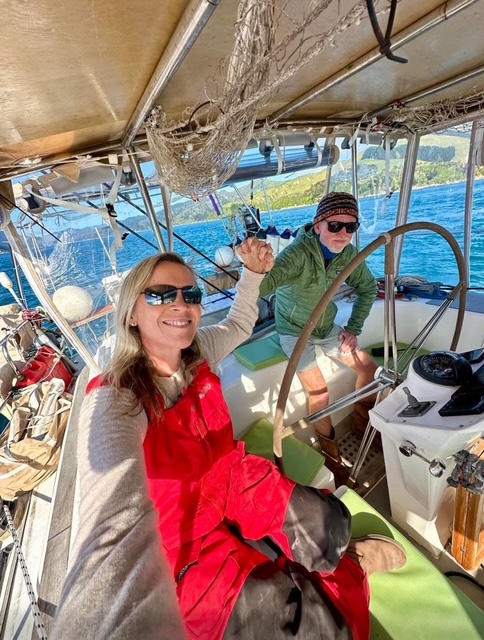 Two people on a boat, smiling and waving, with water and land in the background, under a canopy.