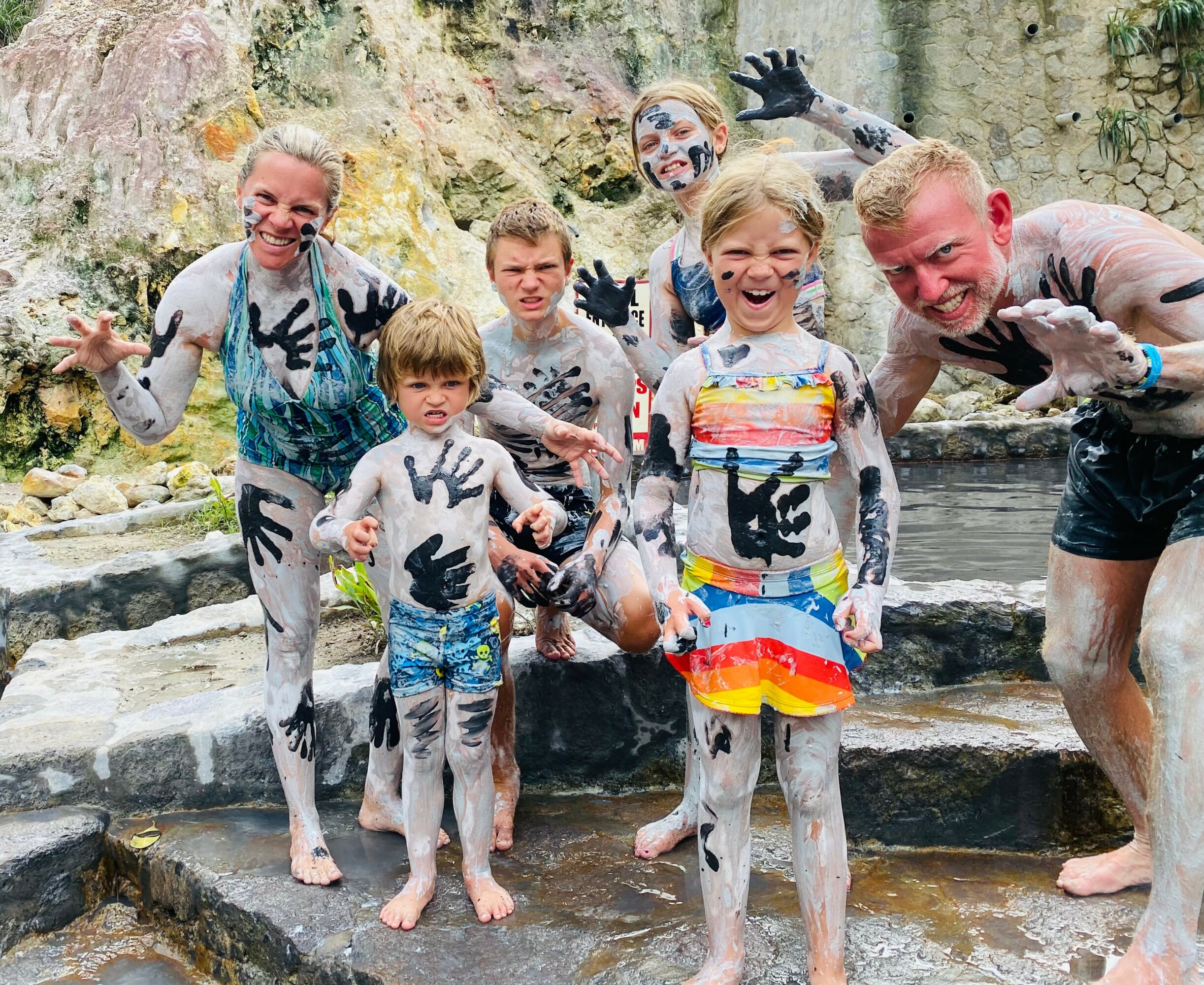 Group of children and adults smiling and playing in a natural outdoor setting, covered in mud or paint, with rocks and water.