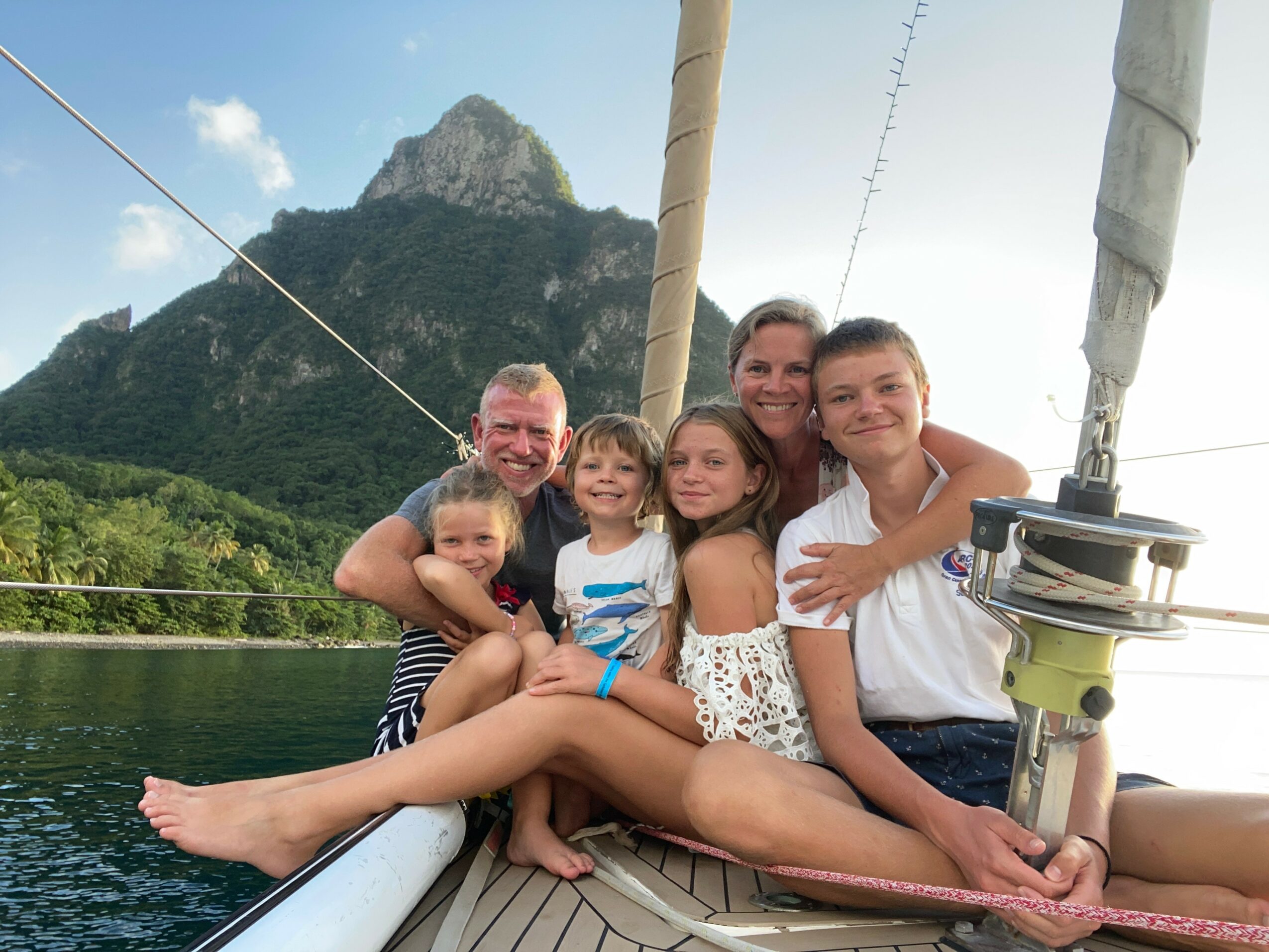 Group of six people sitting on a boat with a mountain and water in the background, smiling and posing together.