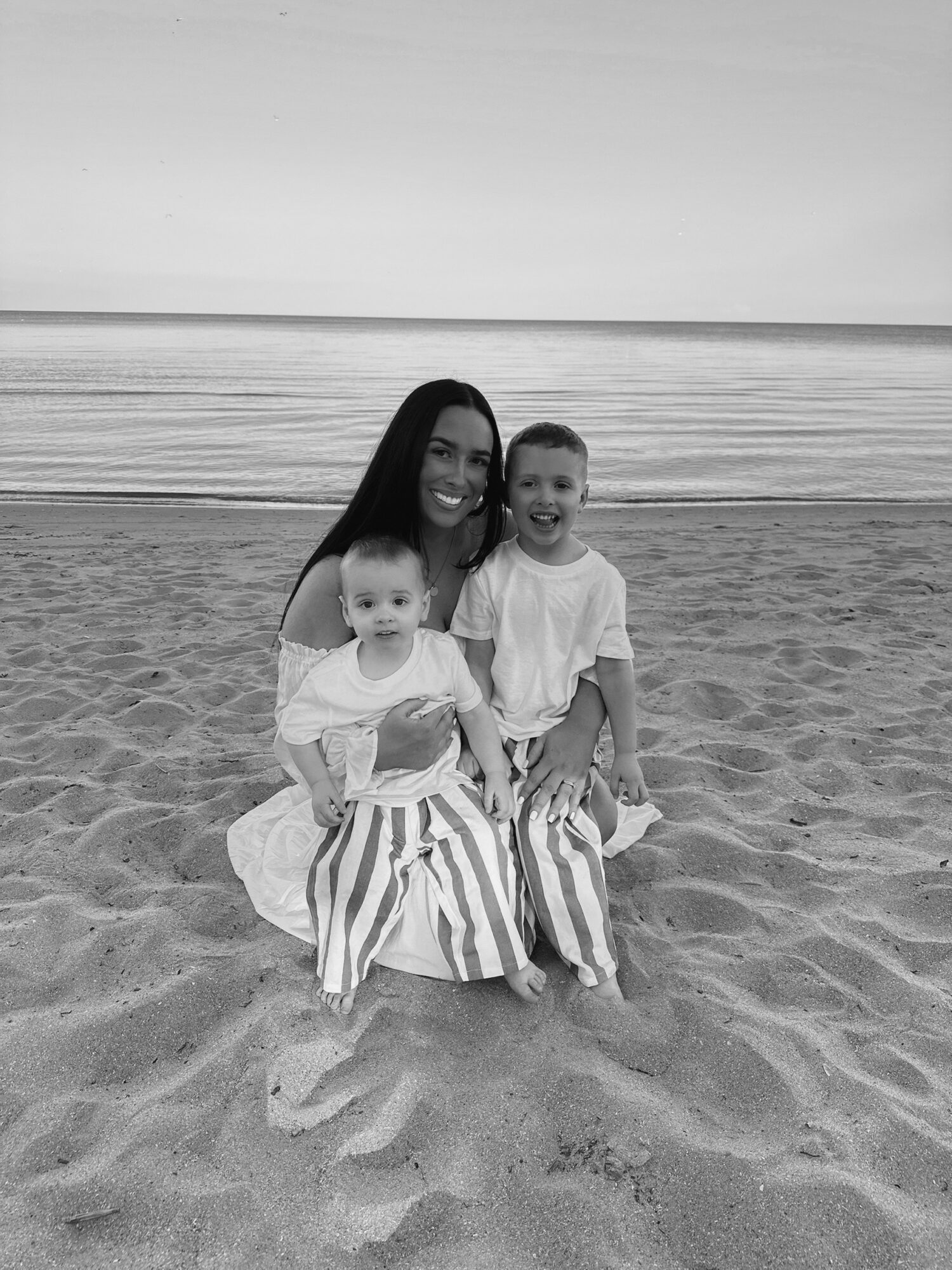 A woman and two children sitting on the beach sand, smiling, with the ocean and sky in the background.