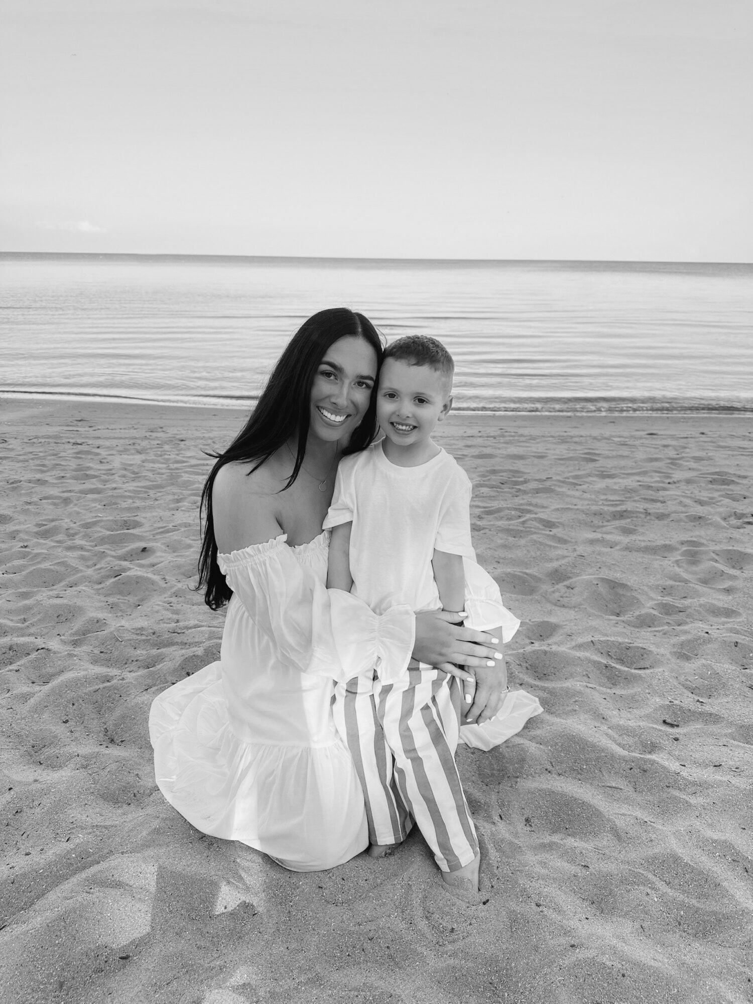 Woman and child sitting on the beach, smiling, with ocean in background, in black and white.