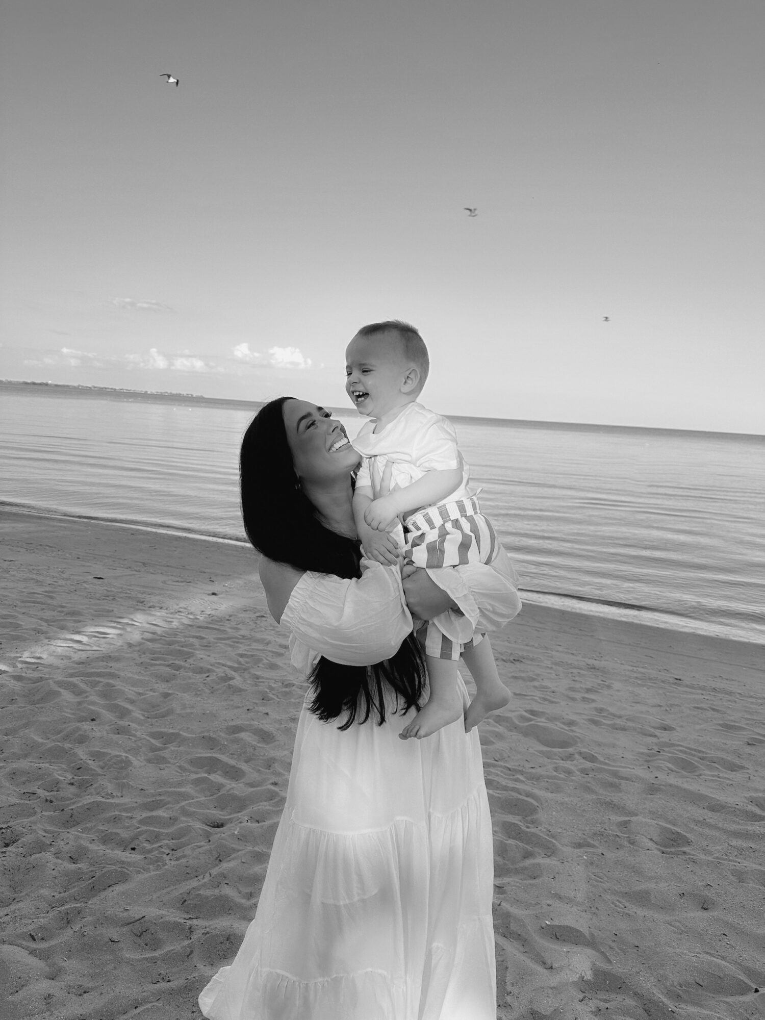 Woman holding a smiling child on a beach with ocean in background, black and white photo.