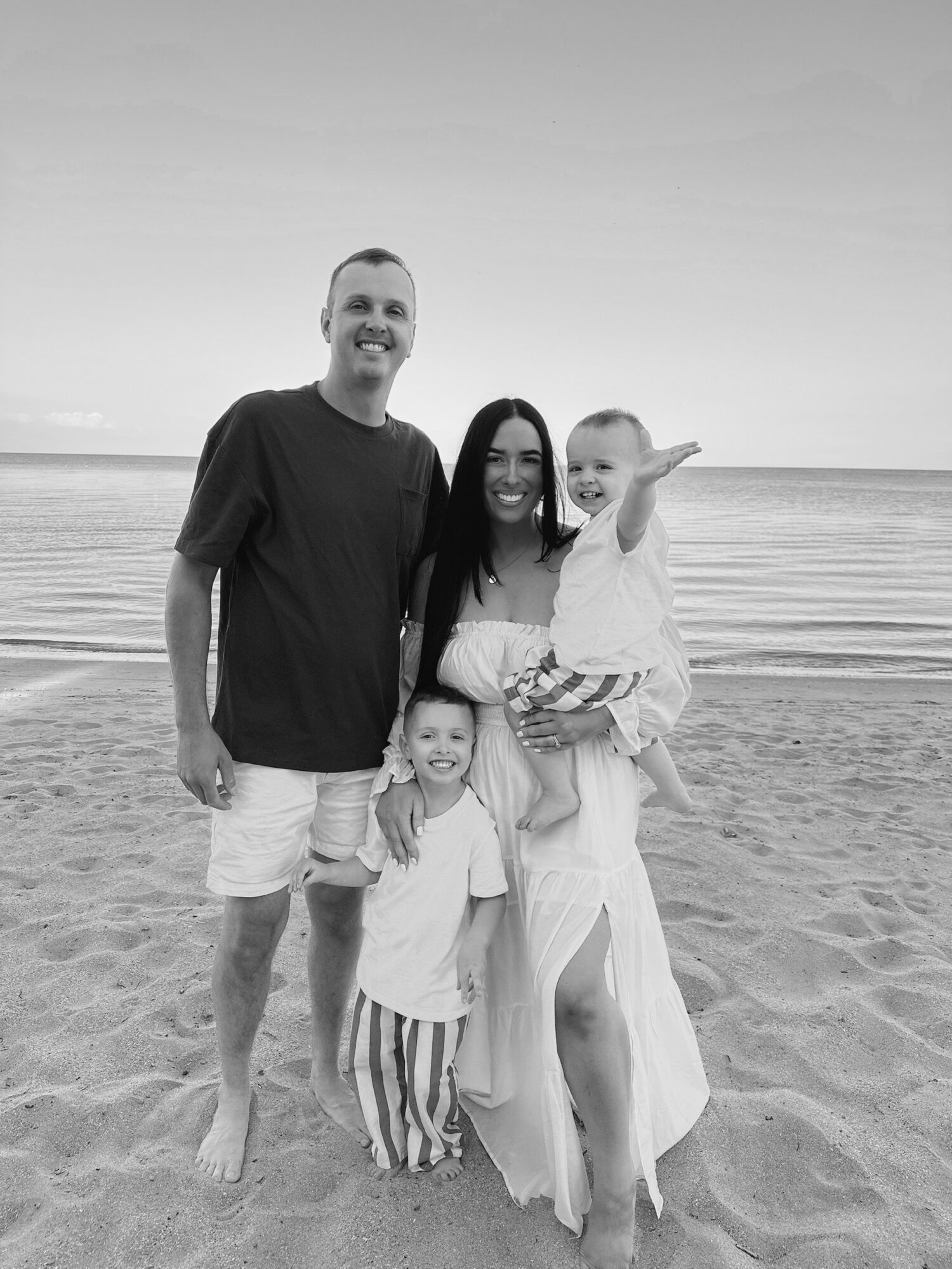 Family of four on the beach, smiling, with the ocean in the background, two adults and two children, one child being held.