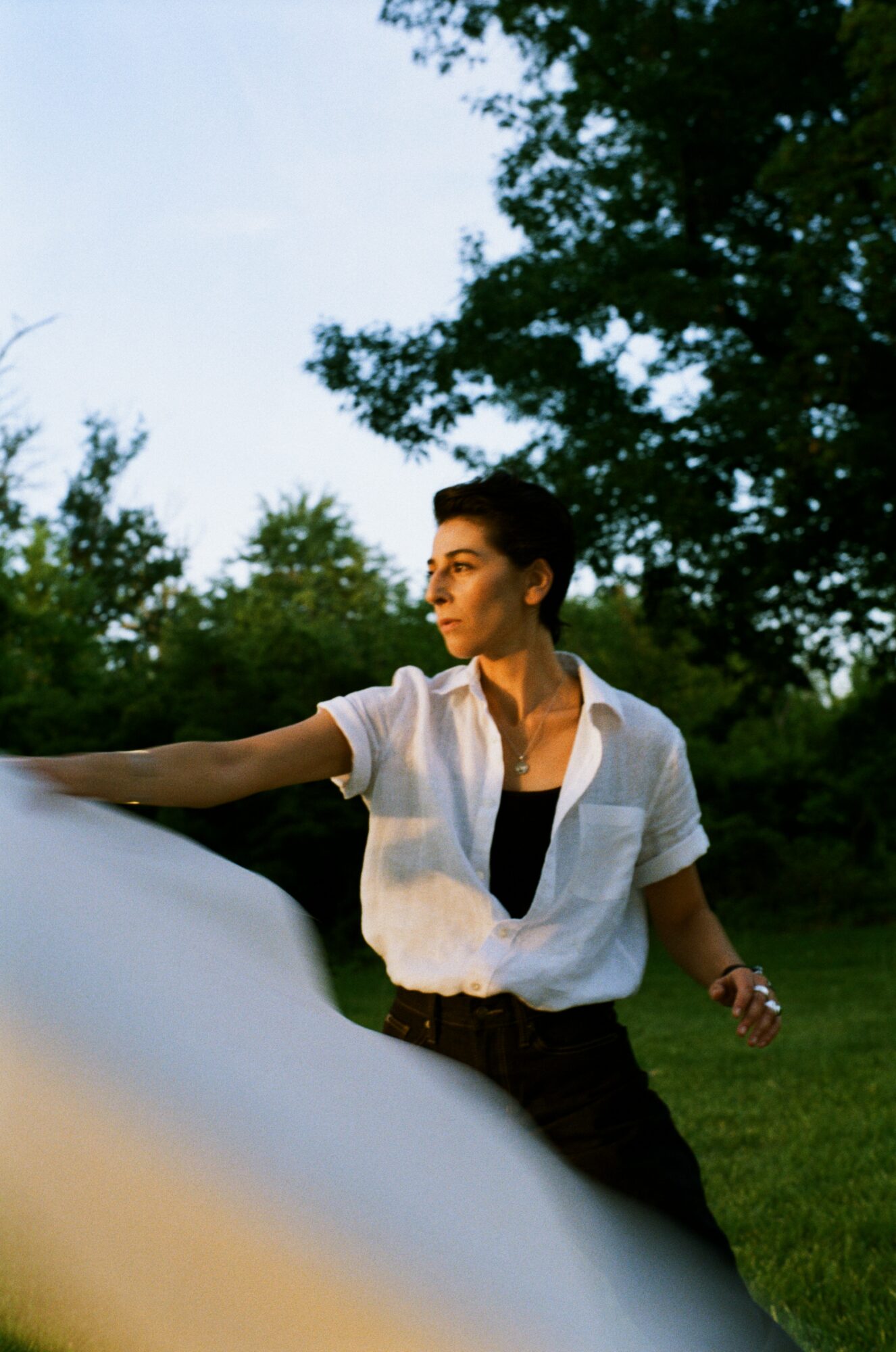 Person with short dark hair in a white shirt outdoors, reaching out with arm extended, trees in background.