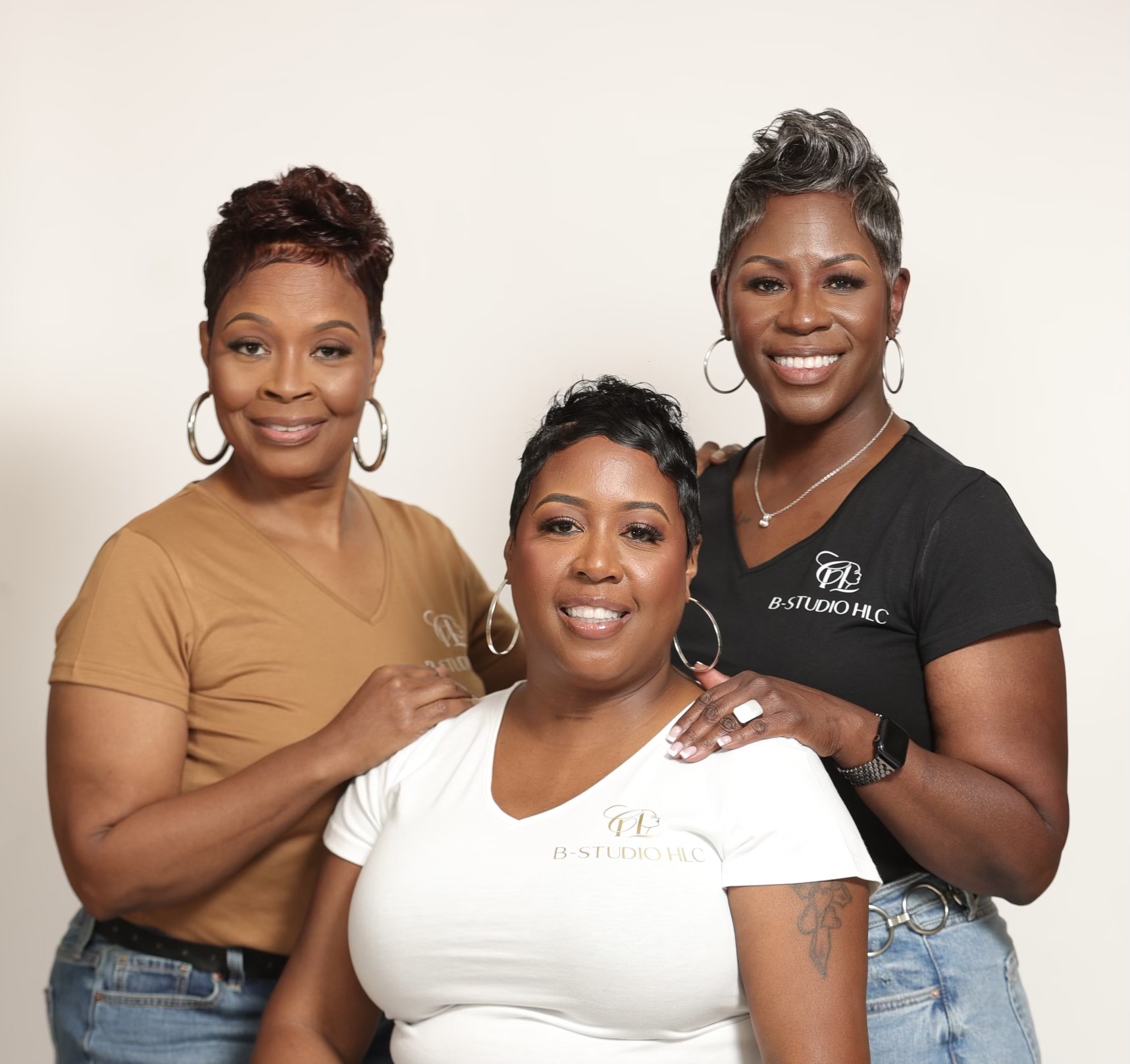 Three women smiling, standing close together, one seated, against a plain light background.