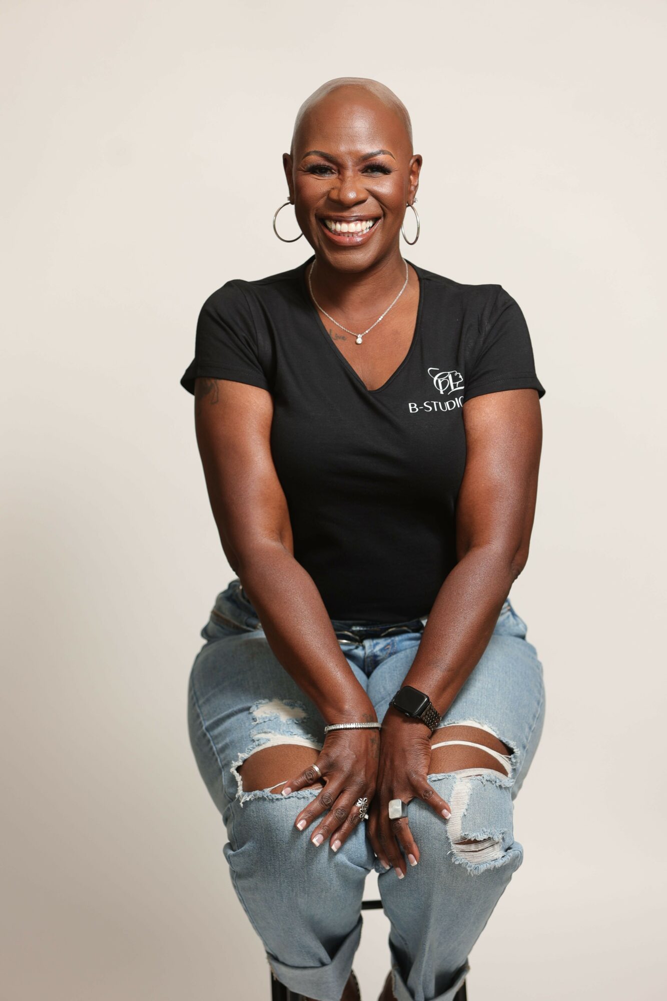 Smiling woman with short hair, wearing a black t-shirt and ripped jeans, sitting against a plain background.