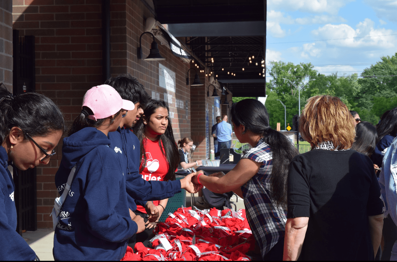 Group of people gathered outside a building, some receiving items from a woman behind a table with red bags, sunny day.