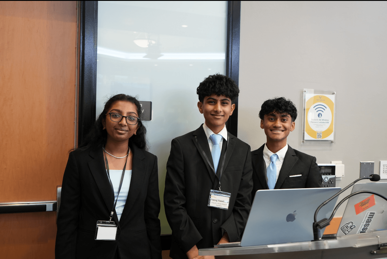 Three young people in formal attire standing behind a laptop in a conference room.
