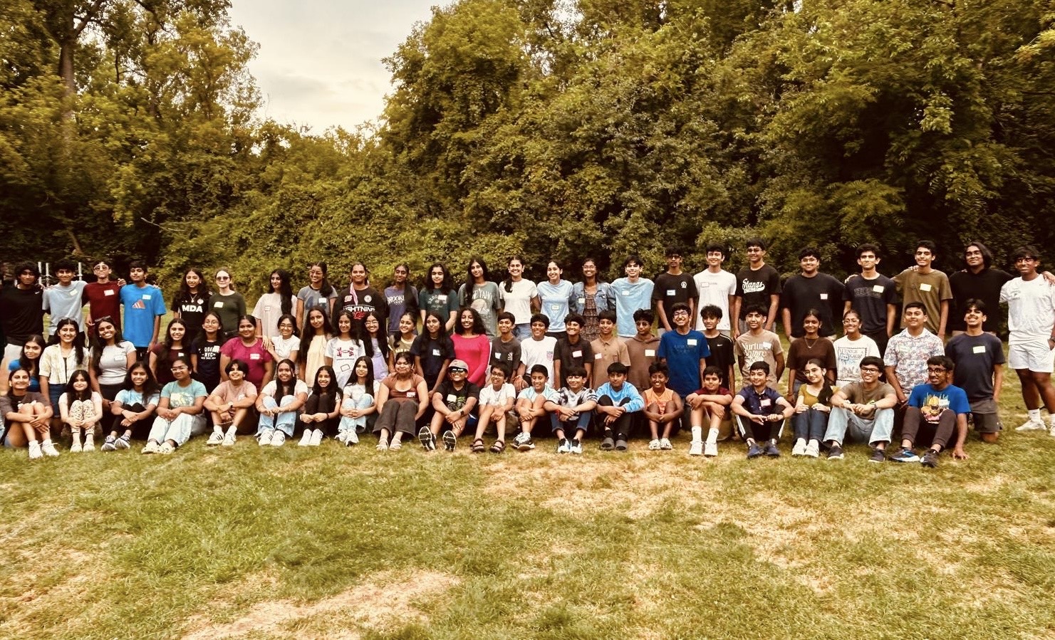 Large group of people outdoors on grass with trees in background, some standing and some sitting, in a park setting.