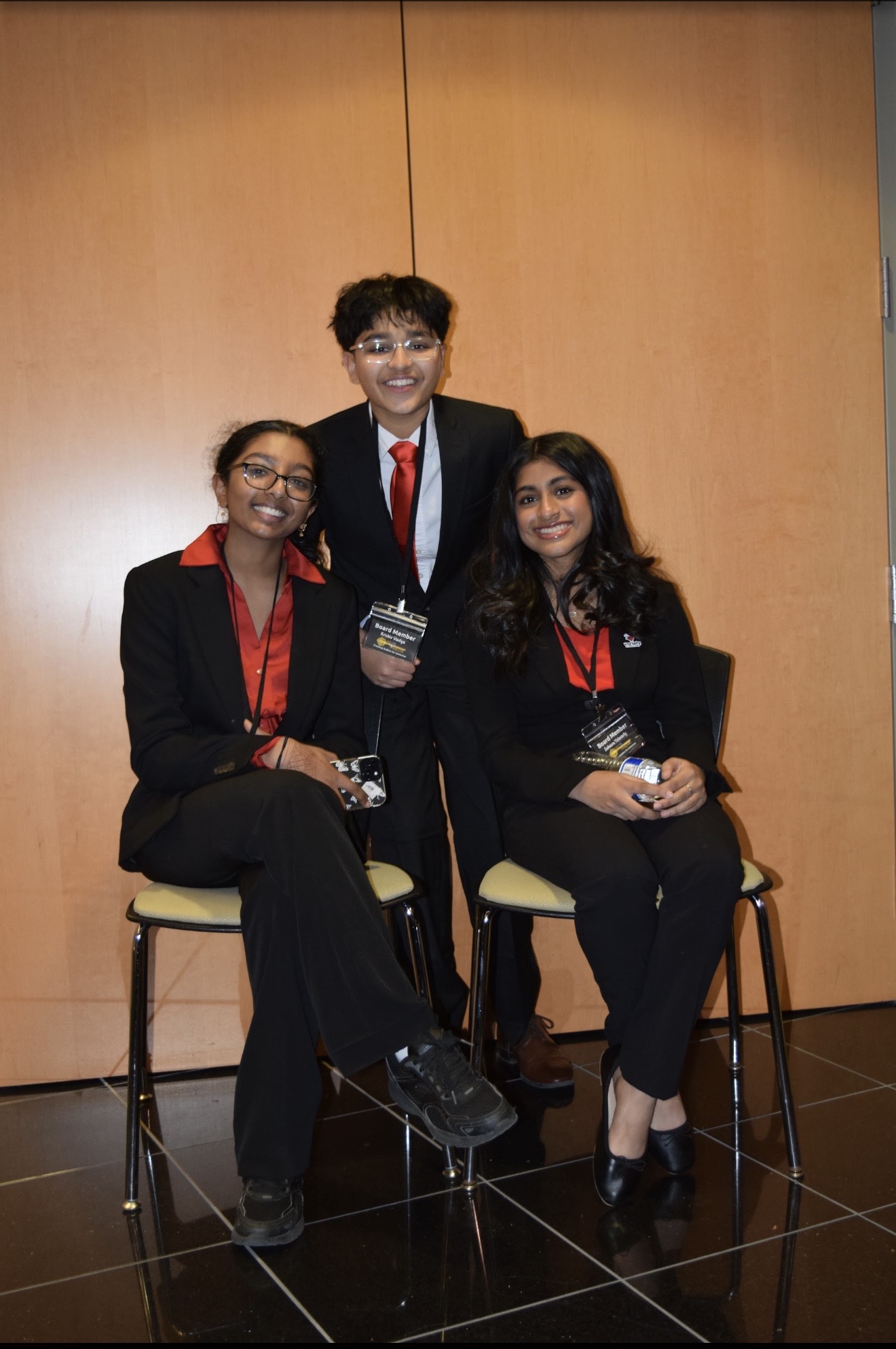 Three people in formal attire posing together, two seated and one standing, in front of a wooden wall.
