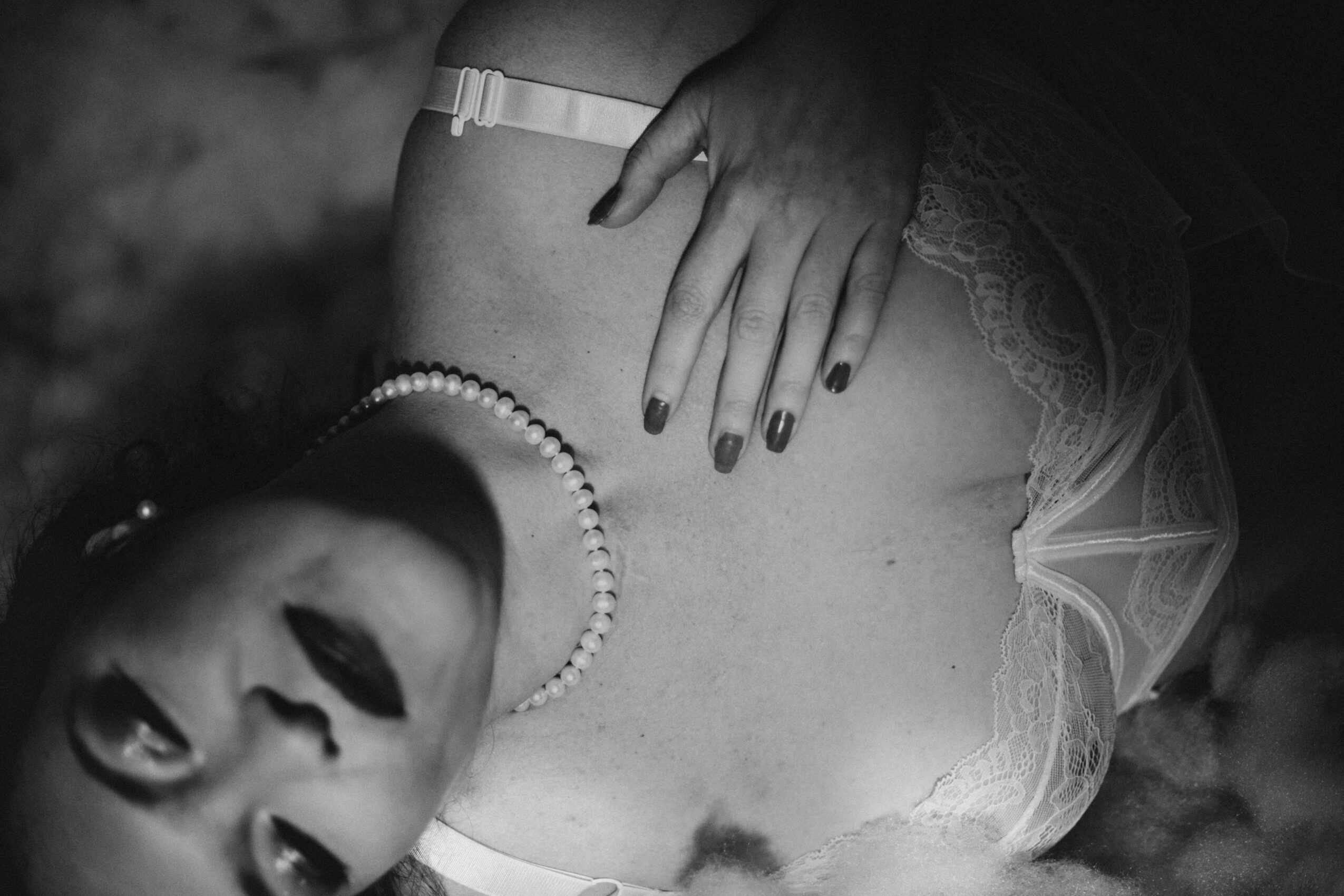 Black and white photo of a woman lying down, wearing a pearl necklace and lace lingerie, with her hand on her chest.