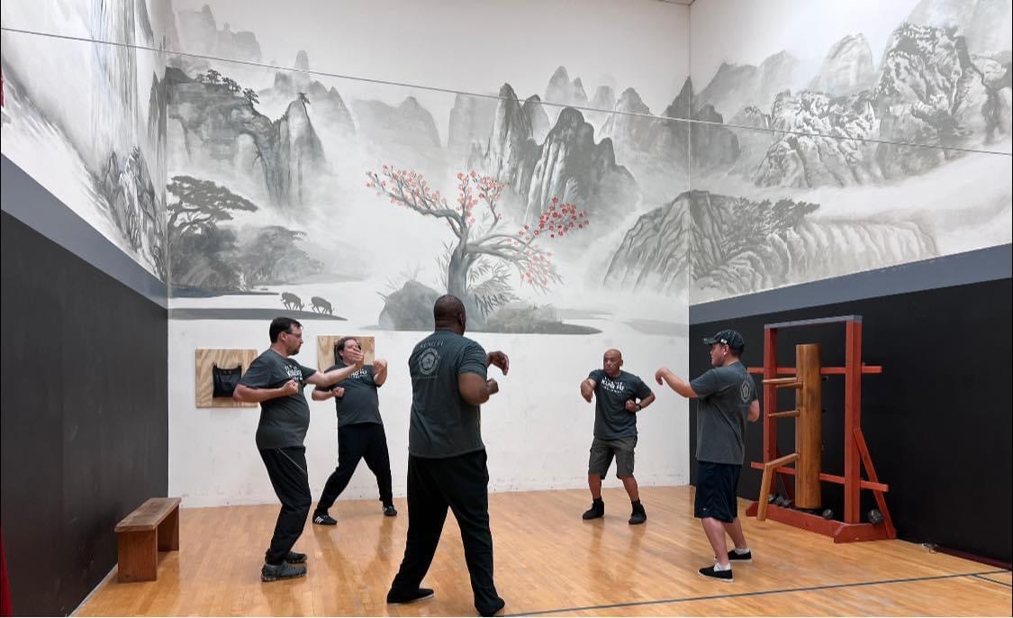 Six people practicing martial arts in a room with a large traditional landscape painting on the wall.