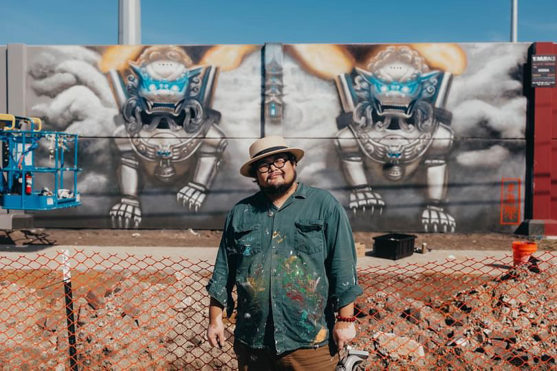 Man wearing glasses and a hat stands in front of a mural of two robotic lions on a wall.