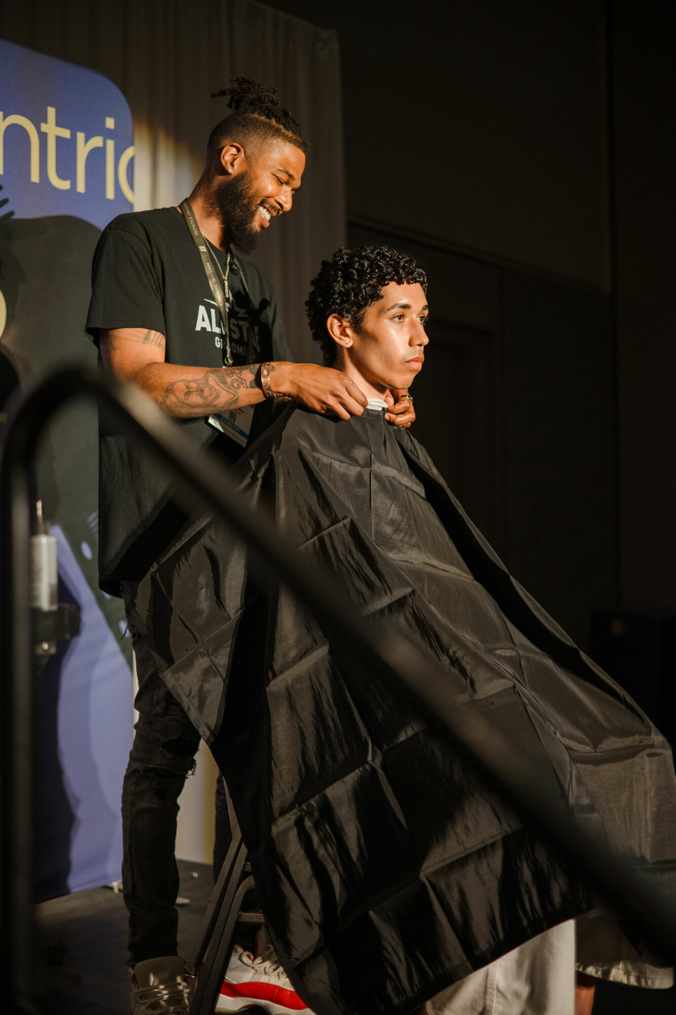 Barber cutting a person's hair on stage, both wearing black capes, with a dark background and partial text visible.