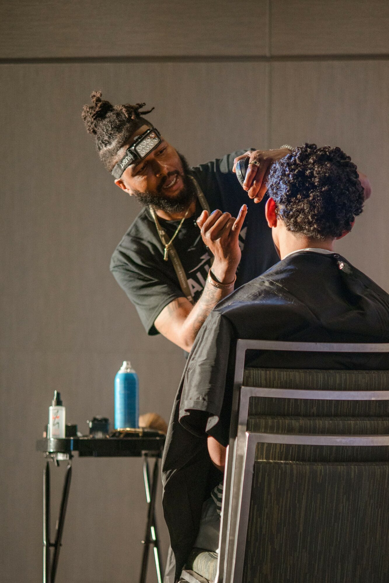 Hairdresser with glasses and a bun styles a person's curly hair in a salon, with hair products on a table nearby.
