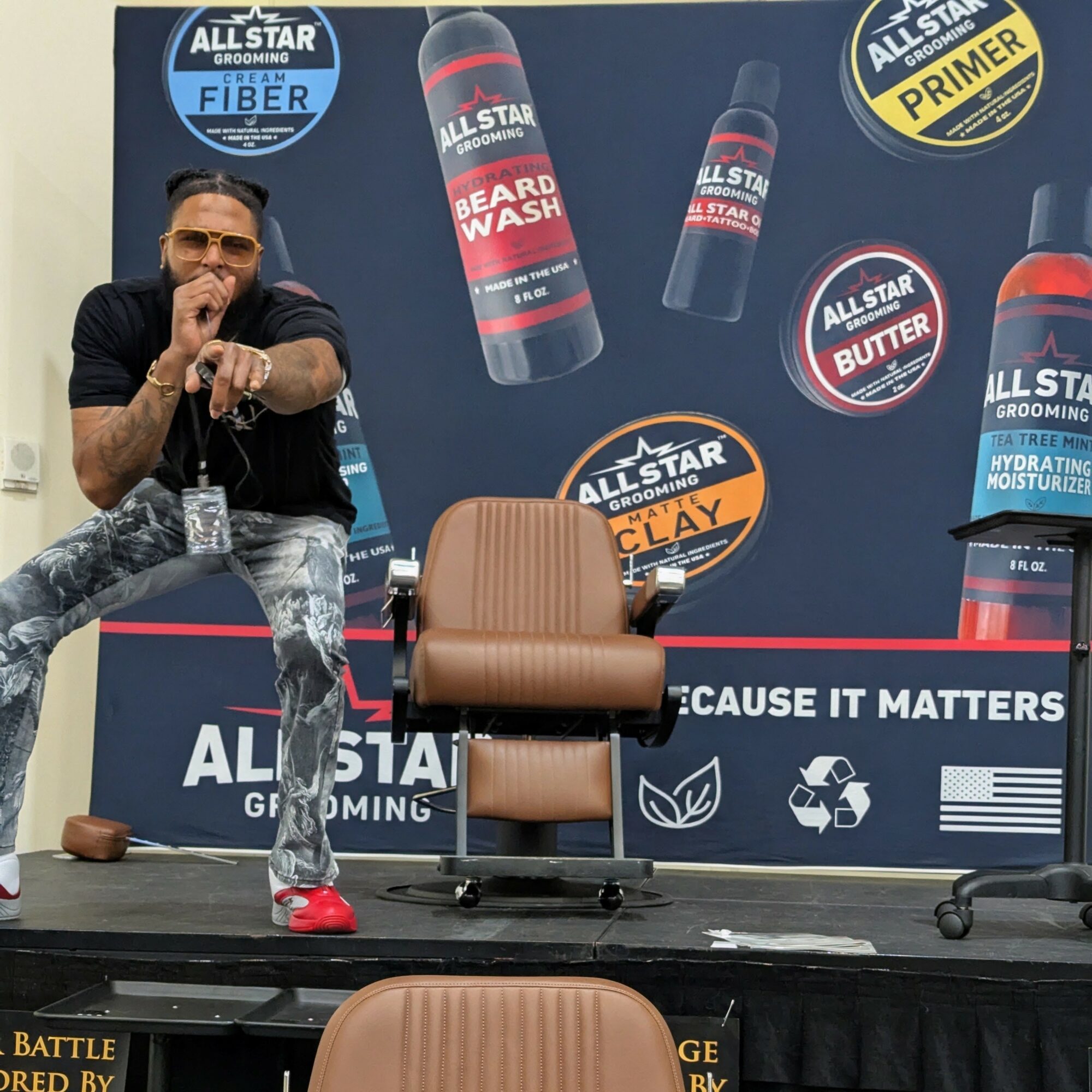 Man sitting on stage with a backdrop displaying hair care products and logos, brown chair in front, stage setup visible.