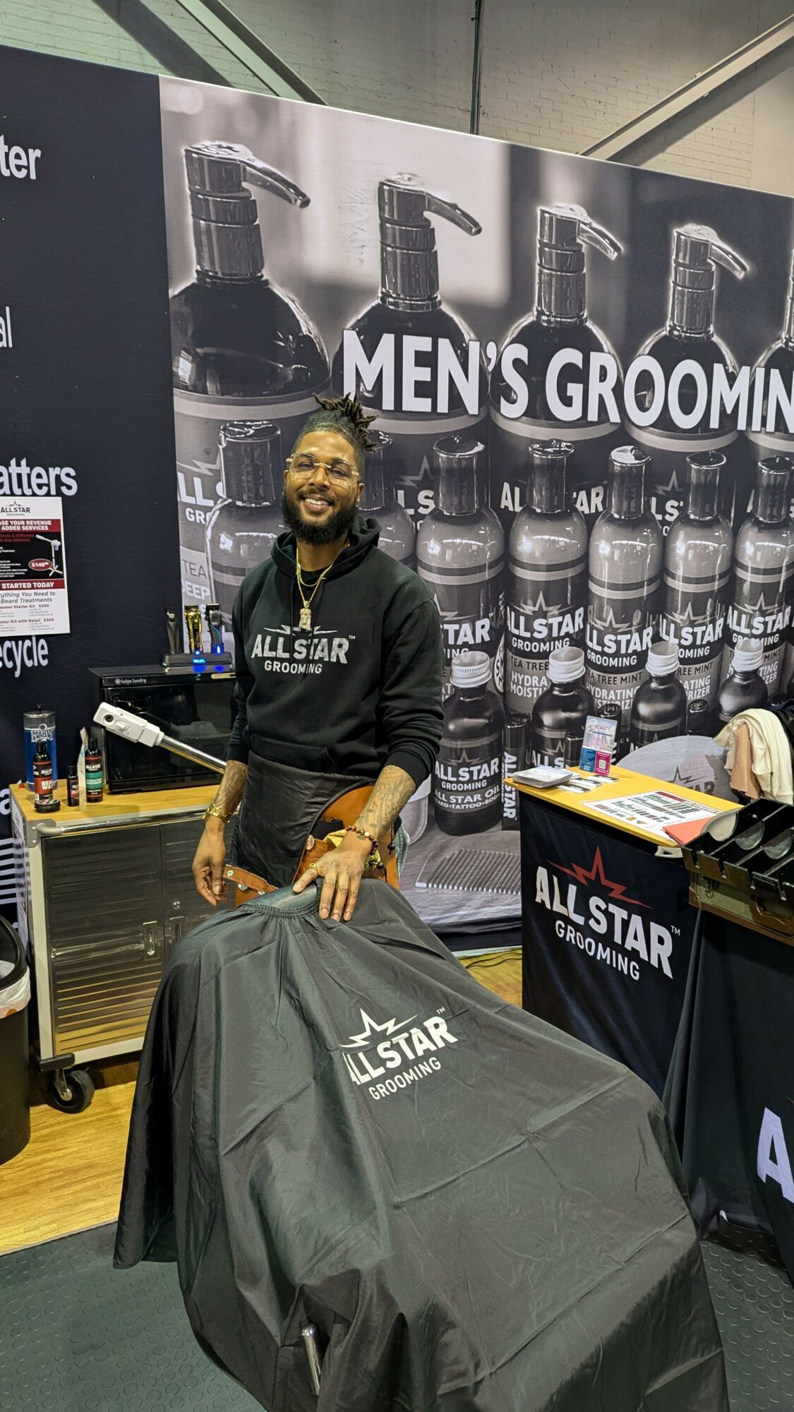 Smiling man standing behind a table with grooming products, in front of a large display of men's grooming spray bottles, at an event.