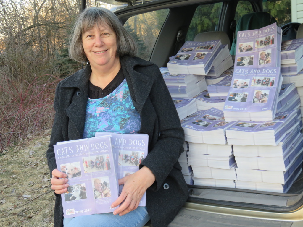 Woman holding a magazine in front of a car filled with stacks of magazines outdoors.