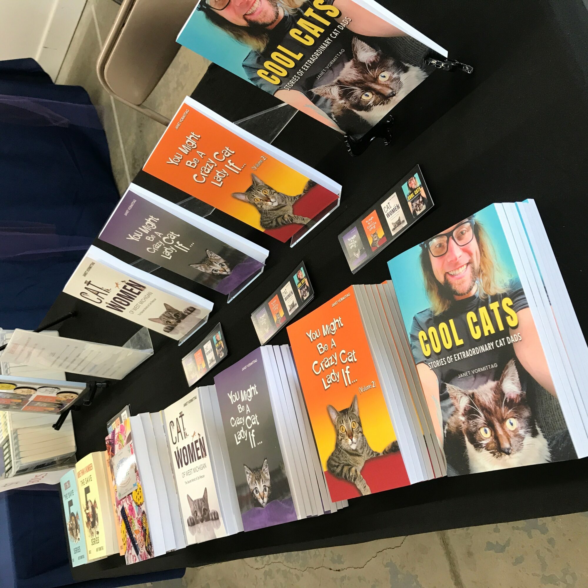 Display of various books about cats, including titles and images of cats and people, arranged on a black table.