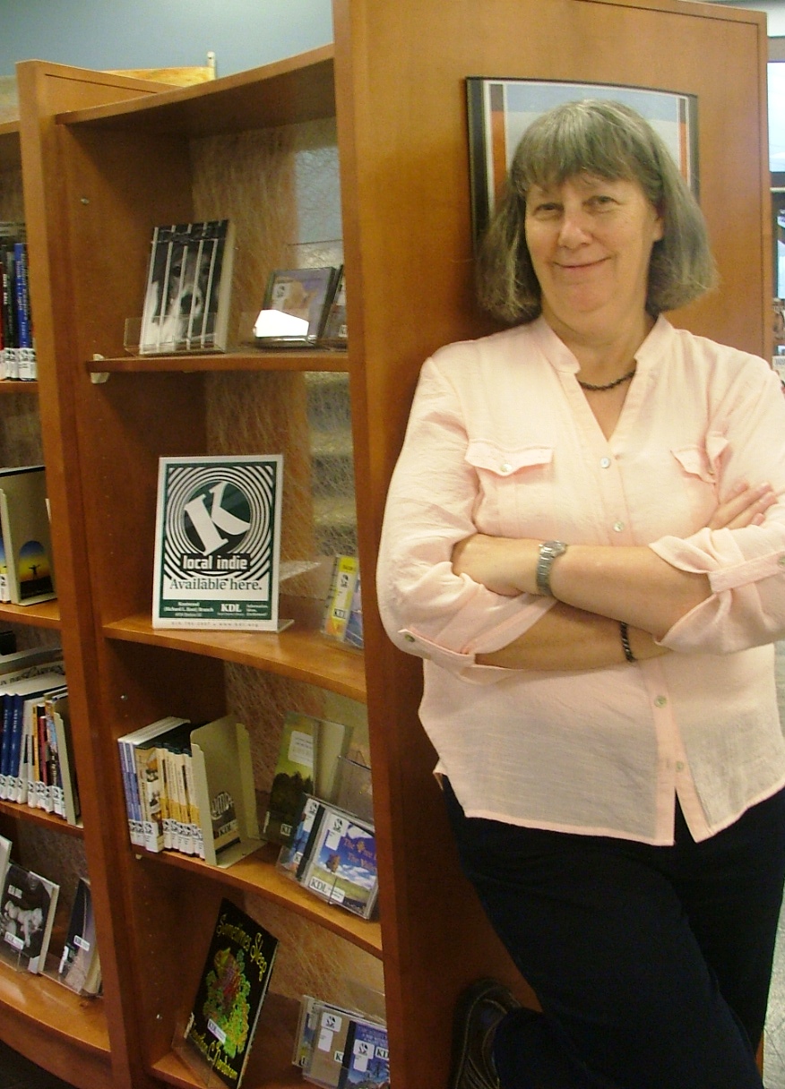 Woman with shoulder-length hair, wearing a light pink shirt and black pants, standing with arms crossed next to a wooden bookshelf.
