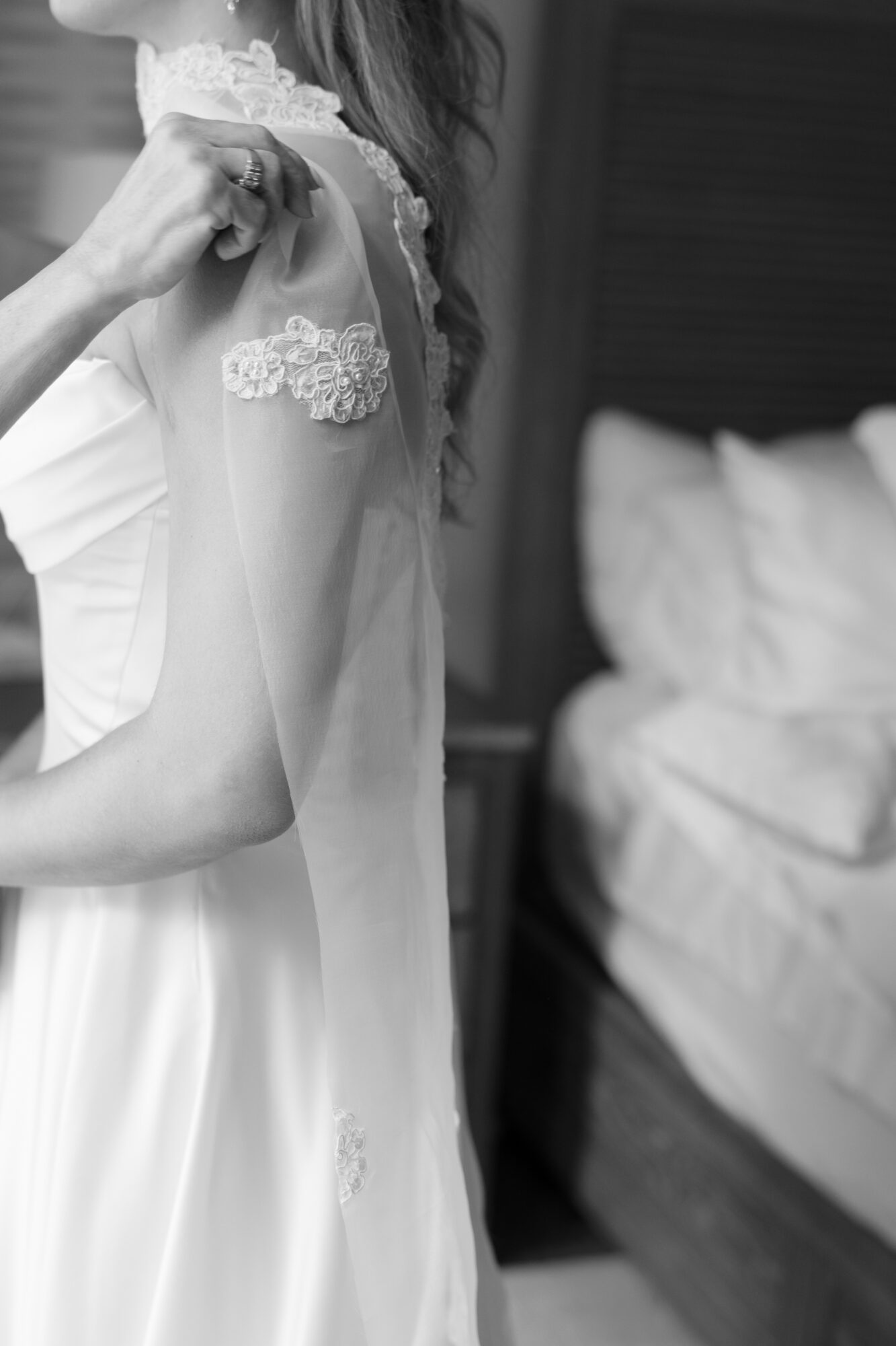 Close-up of a woman in a dress with lace details on the shoulder, standing near a bed in a bedroom.