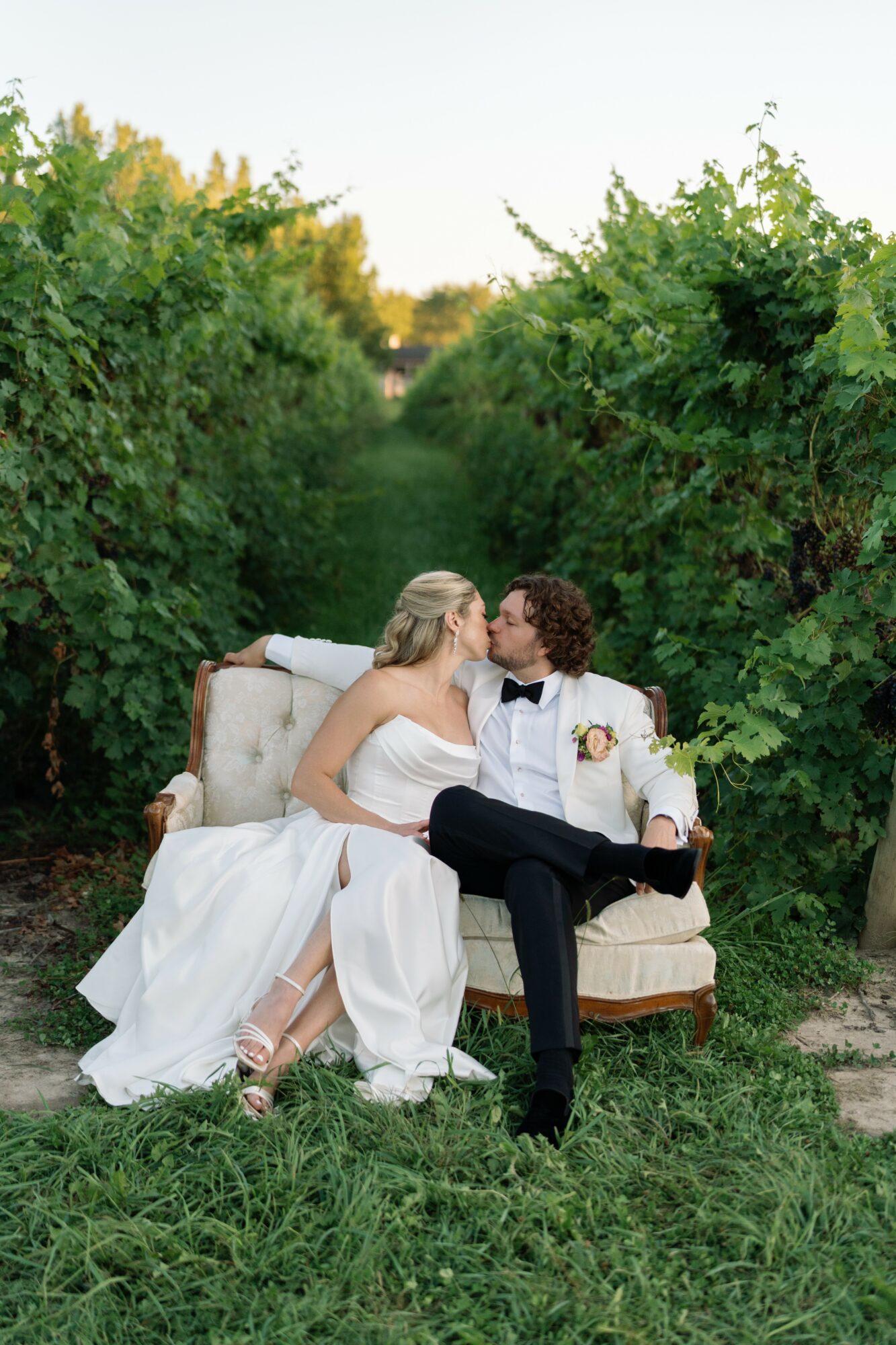 Bride and groom sitting on a vintage sofa outdoors, surrounded by green foliage, sharing a kiss.