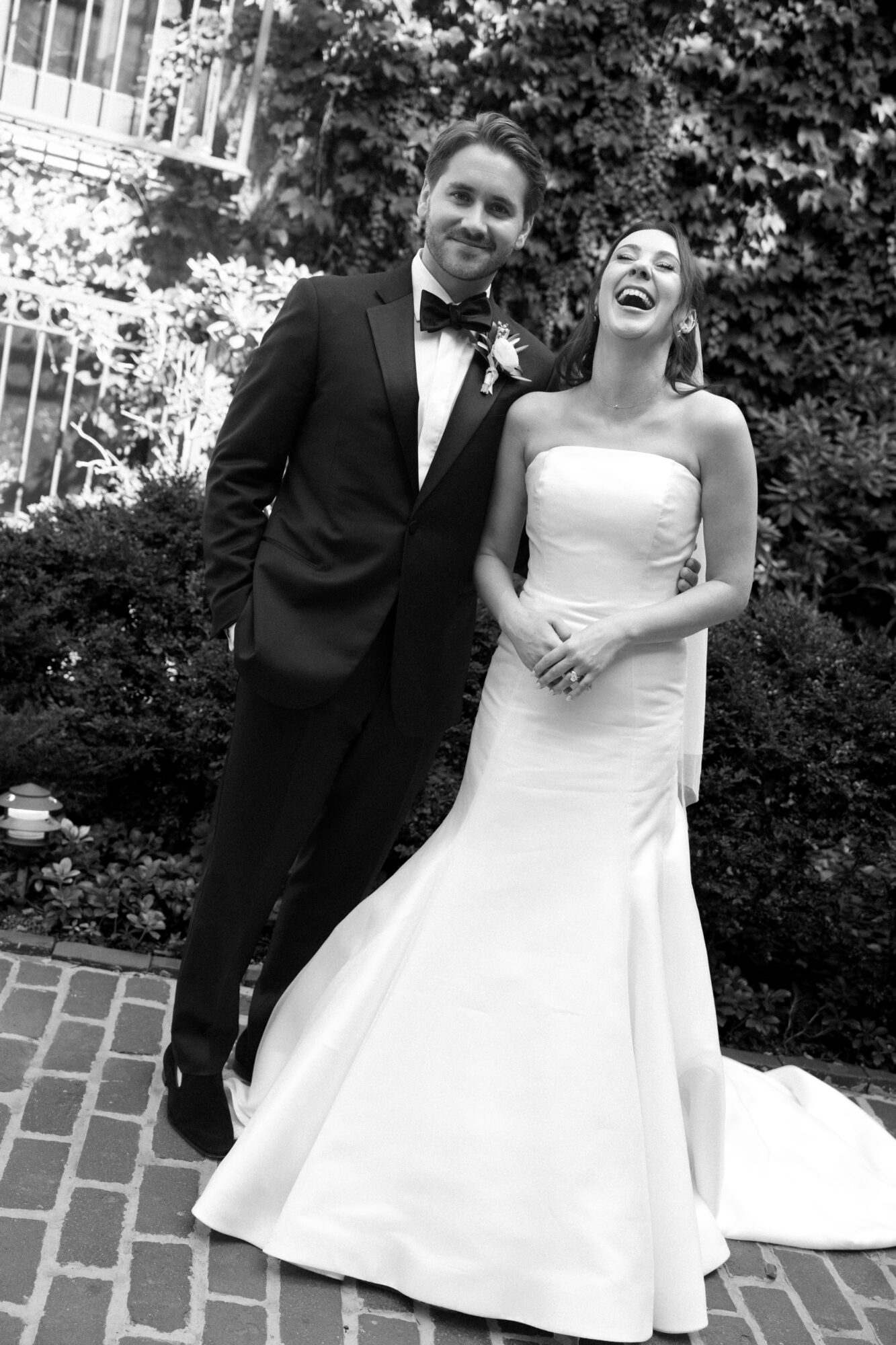 A man in a tuxedo and a woman in a wedding dress stand outdoors, smiling, with greenery and a brick path behind them.