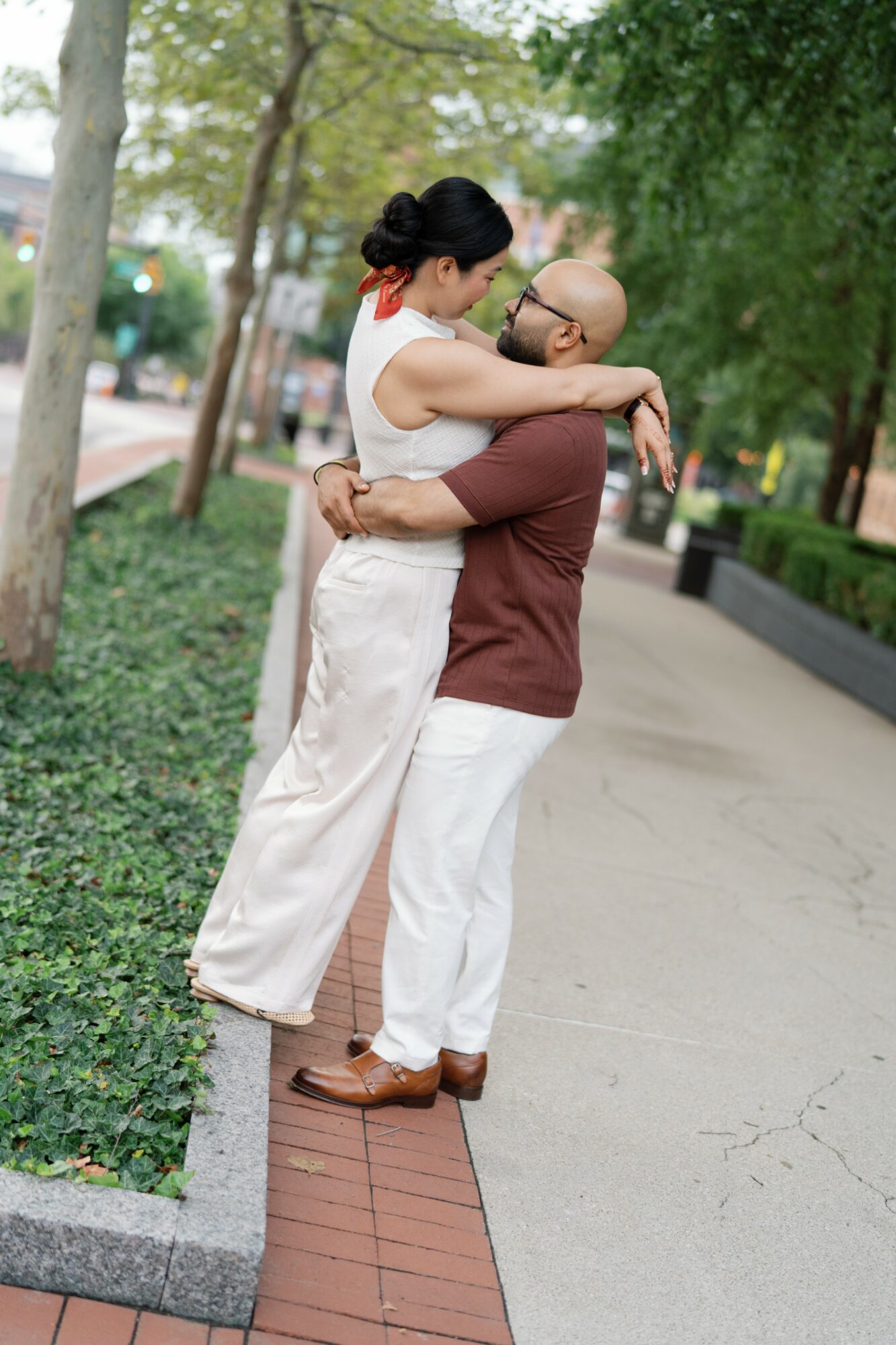 Two women hugging outdoors on a sidewalk with trees and greenery, one with glasses and a bun, the other with dark hair.