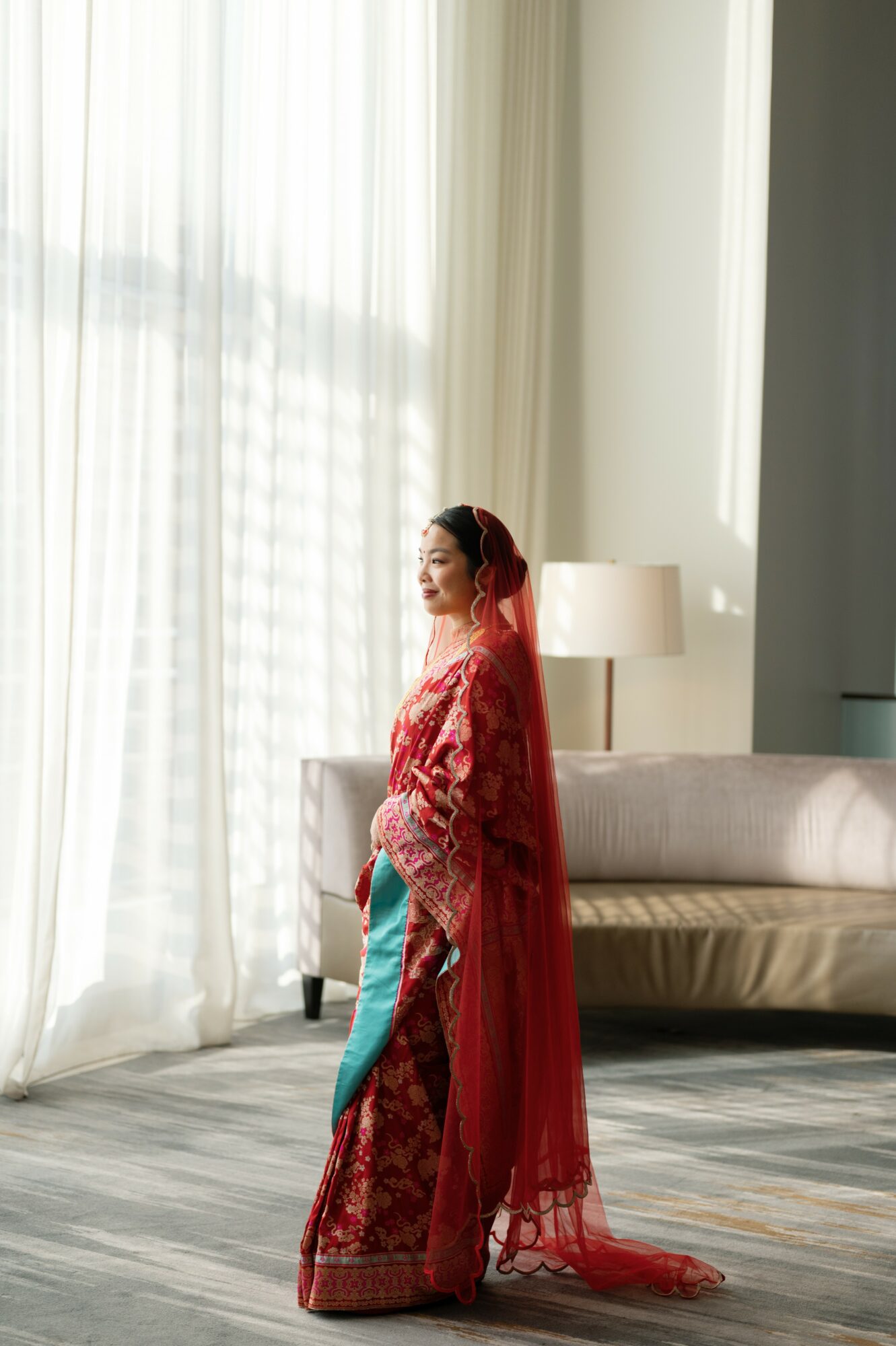 Woman in traditional red attire with a veil, standing in a bright room near a sofa and window.