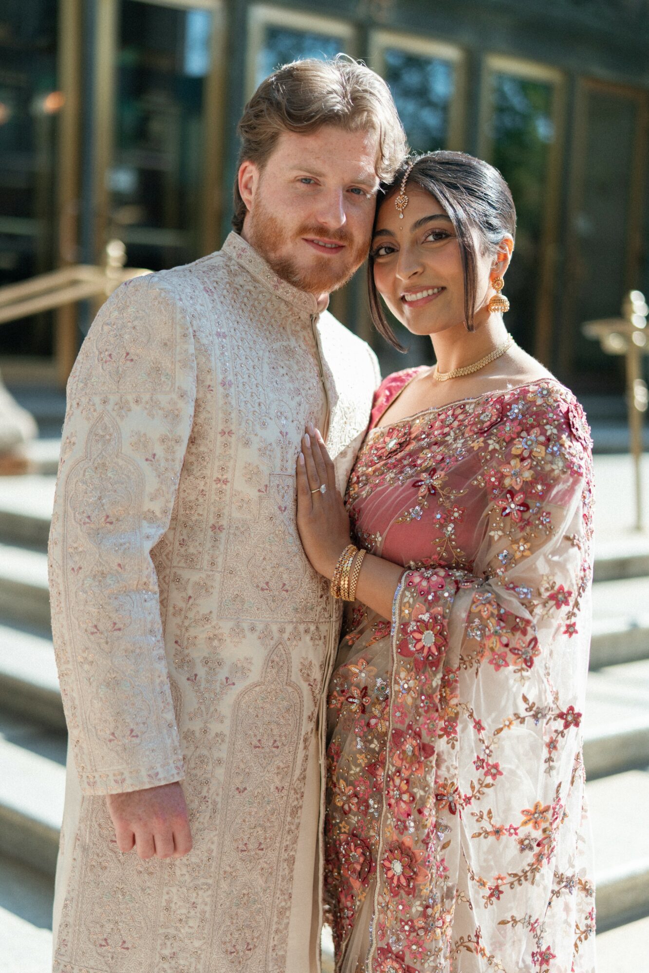 A man and woman in traditional attire standing outdoors, smiling, with a building and glass windows in the background.