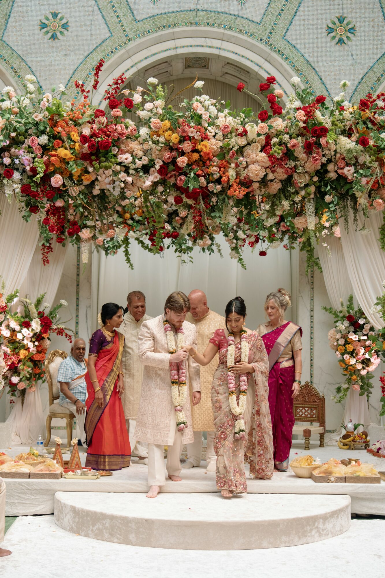 People in traditional attire participate in a wedding ceremony under floral decorations and an ornate arch.