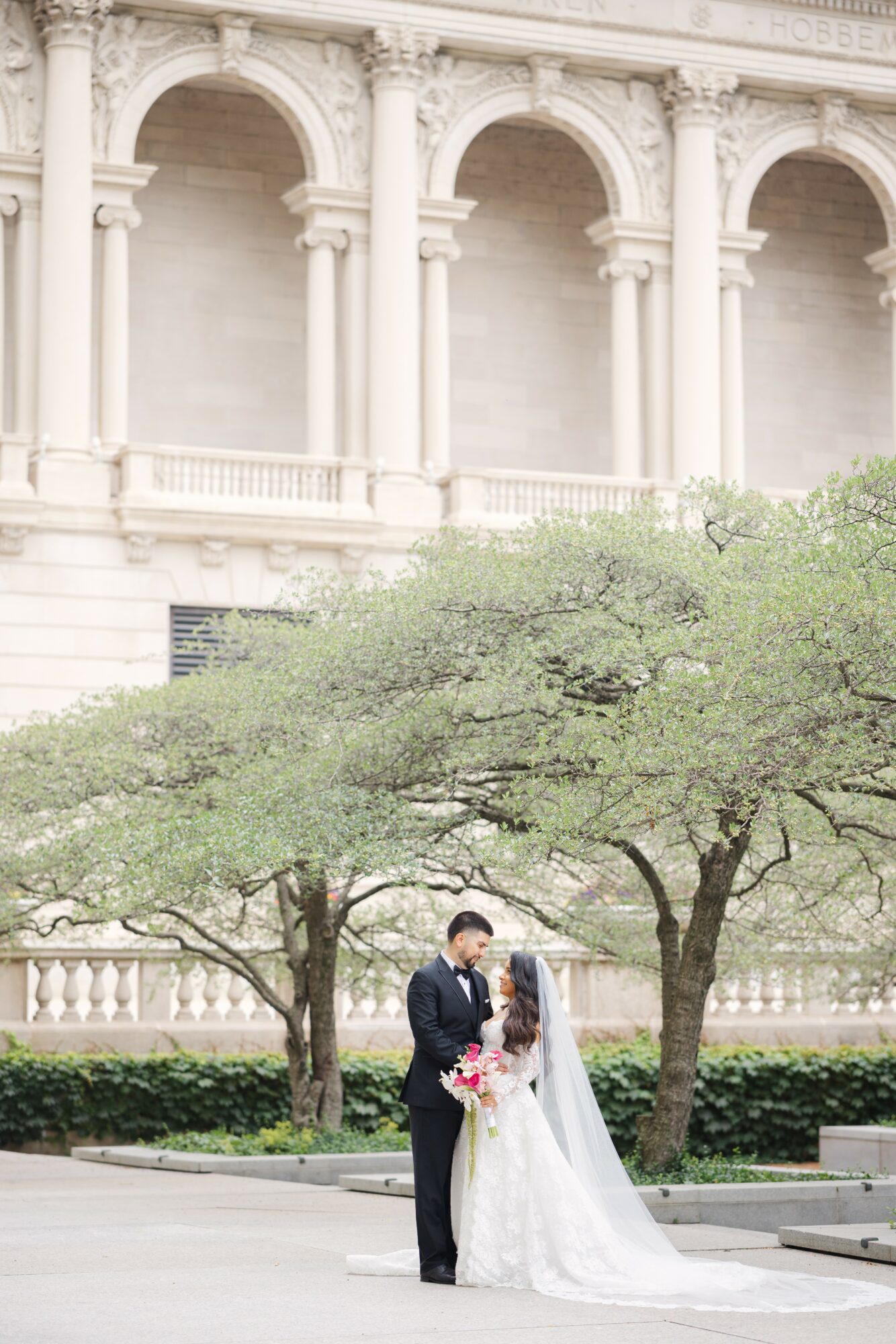 A couple in wedding attire standing under a tree in front of a historic building with arches and columns.